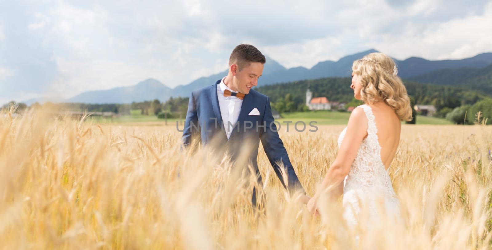 Groom and bride holding hands in wheat field somewhere in Slovenian countryside. by kasto