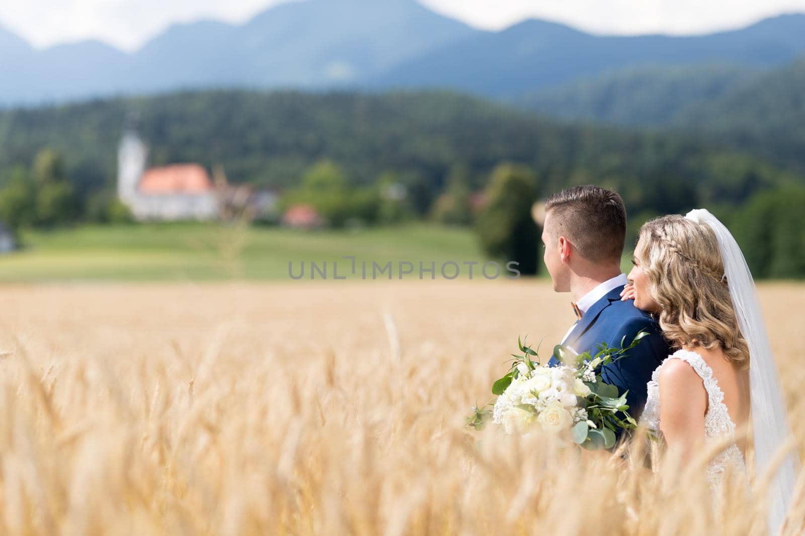 Bride hugs groom tenderly in wheat field somewhere in Slovenian countryside. Caucasian happy romantic young couple celebrating their marriage.