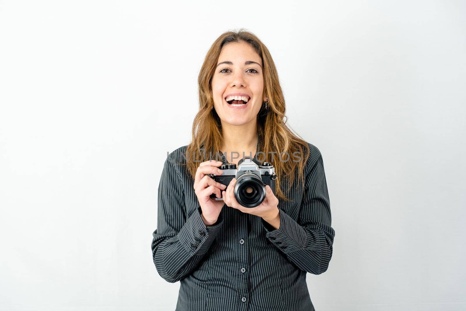 Studio shot of smiling happy young woman holding an old vintage photo camera watching photographer on white background for copy space. Concept of passion for imaging and history of classic photography