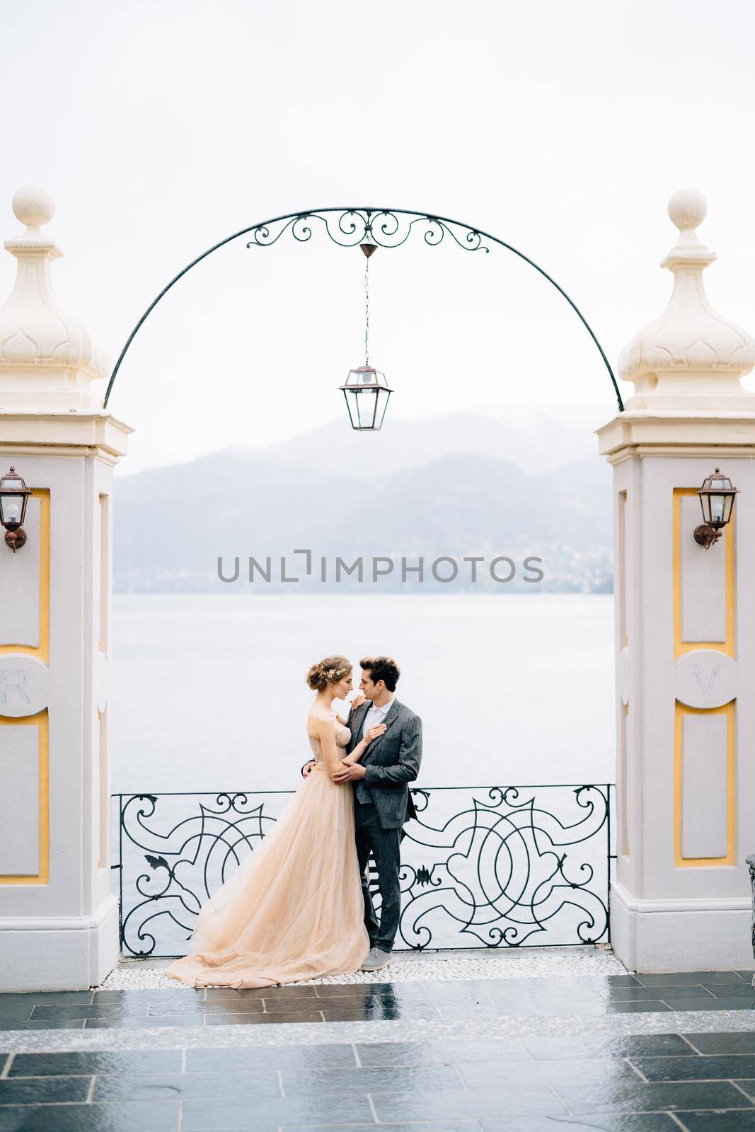 Newlyweds stand in an embrace under an ancient arch leaning on a forged fence against the backdrop of Lake Como. High quality photo