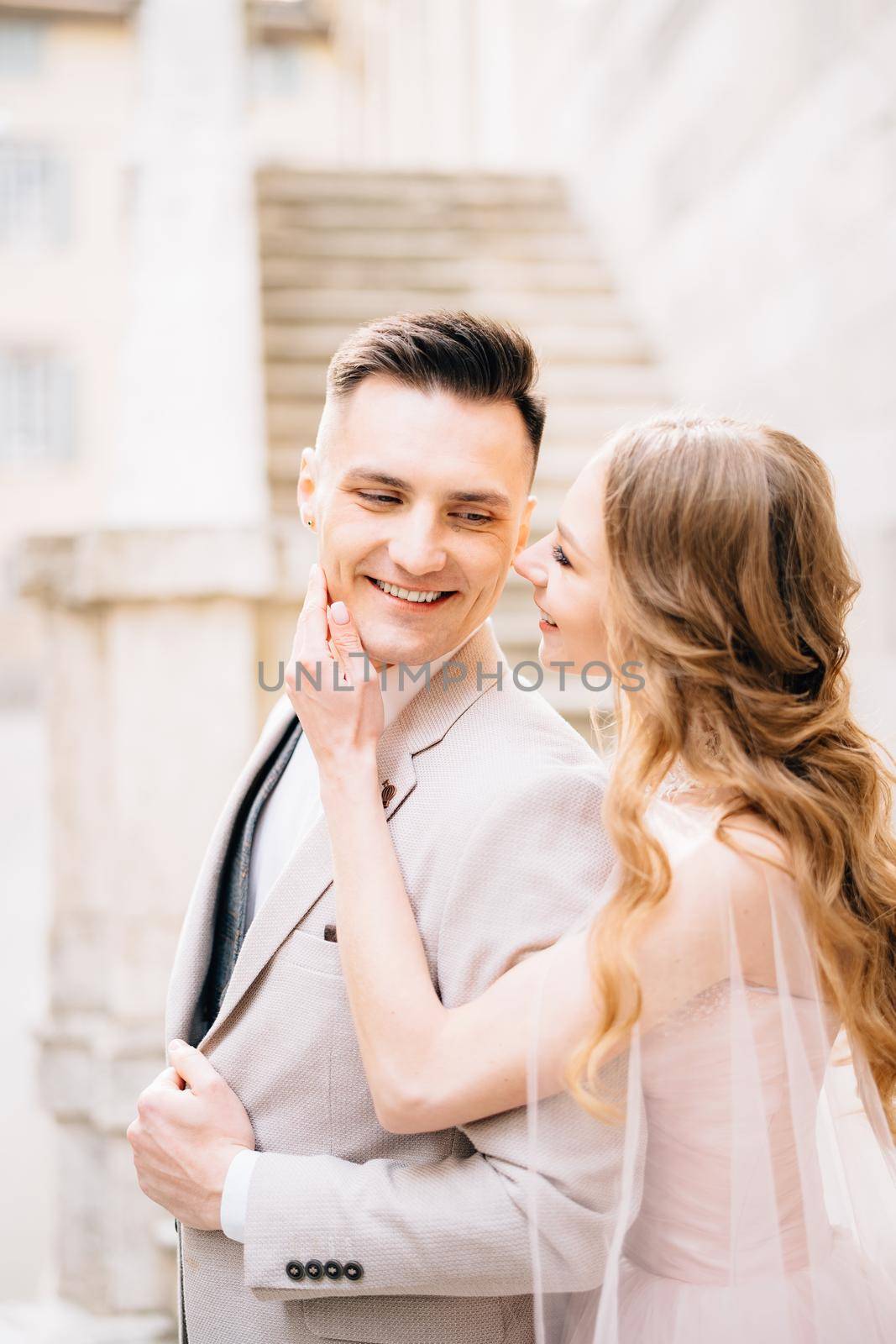 Bride holds her palm on the chin of smiling groom against the background of the steps of an ancient building in Bergamo by Nadtochiy