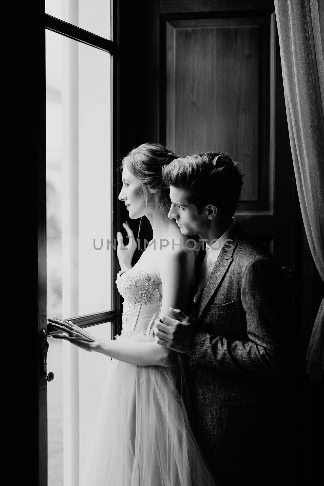 Groom hugs bride from behind at the large window of an old villa. Lake Como. Black and white photo by Nadtochiy