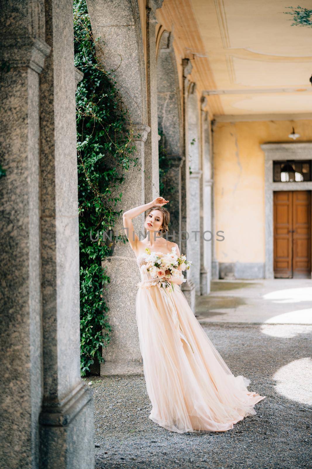 Bride in a beautiful dress with a bouquet of pink flowers stands in the vaulted hall with her hand on her forehead. Lake Como, Italy. High quality photo
