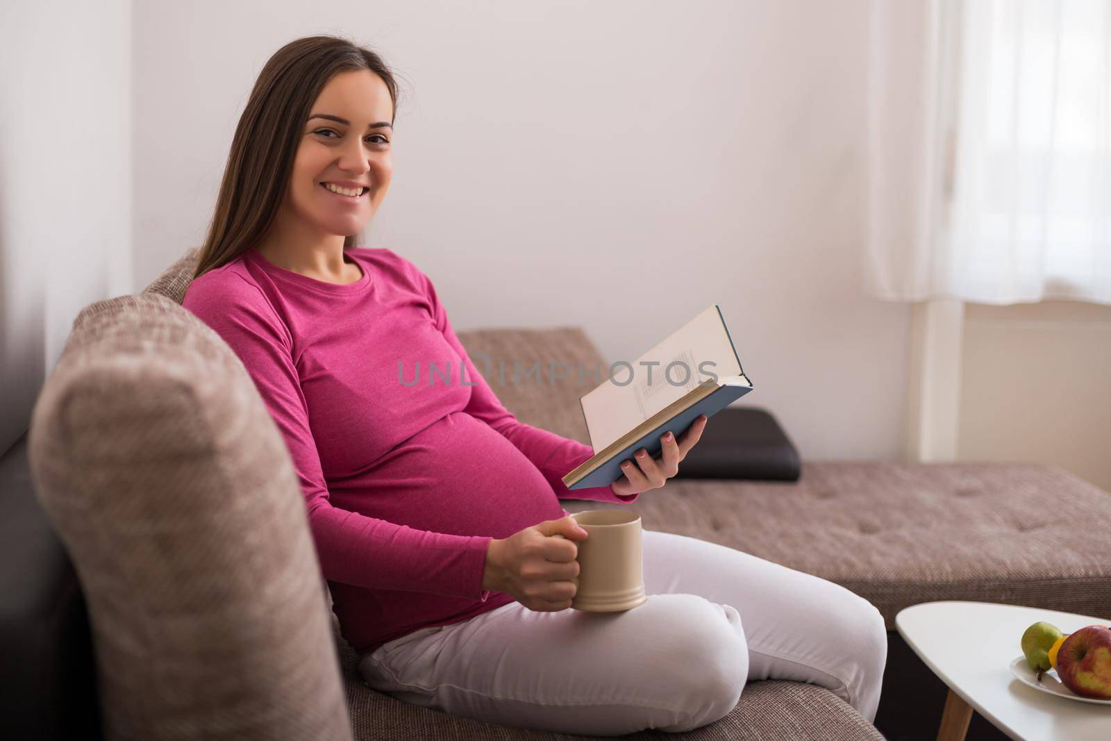 Beautiful pregnant woman enjoys reading book and drinking tea at her home.