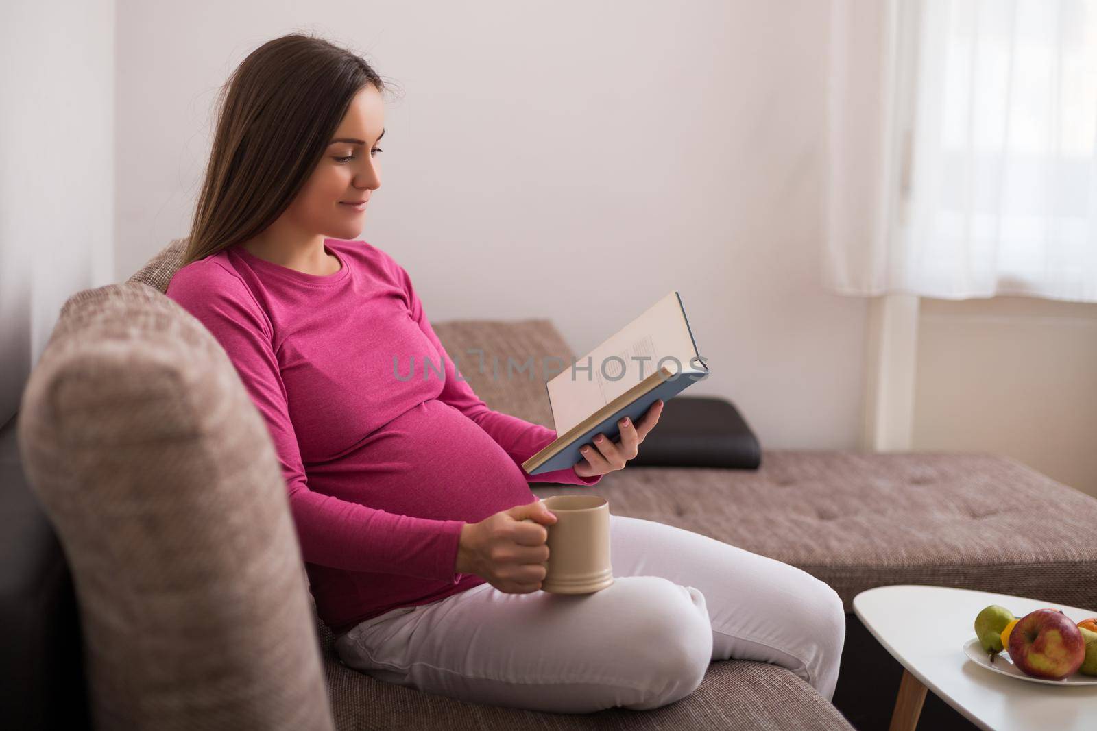 Beautiful pregnant woman enjoys reading book and drinking tea at her home.