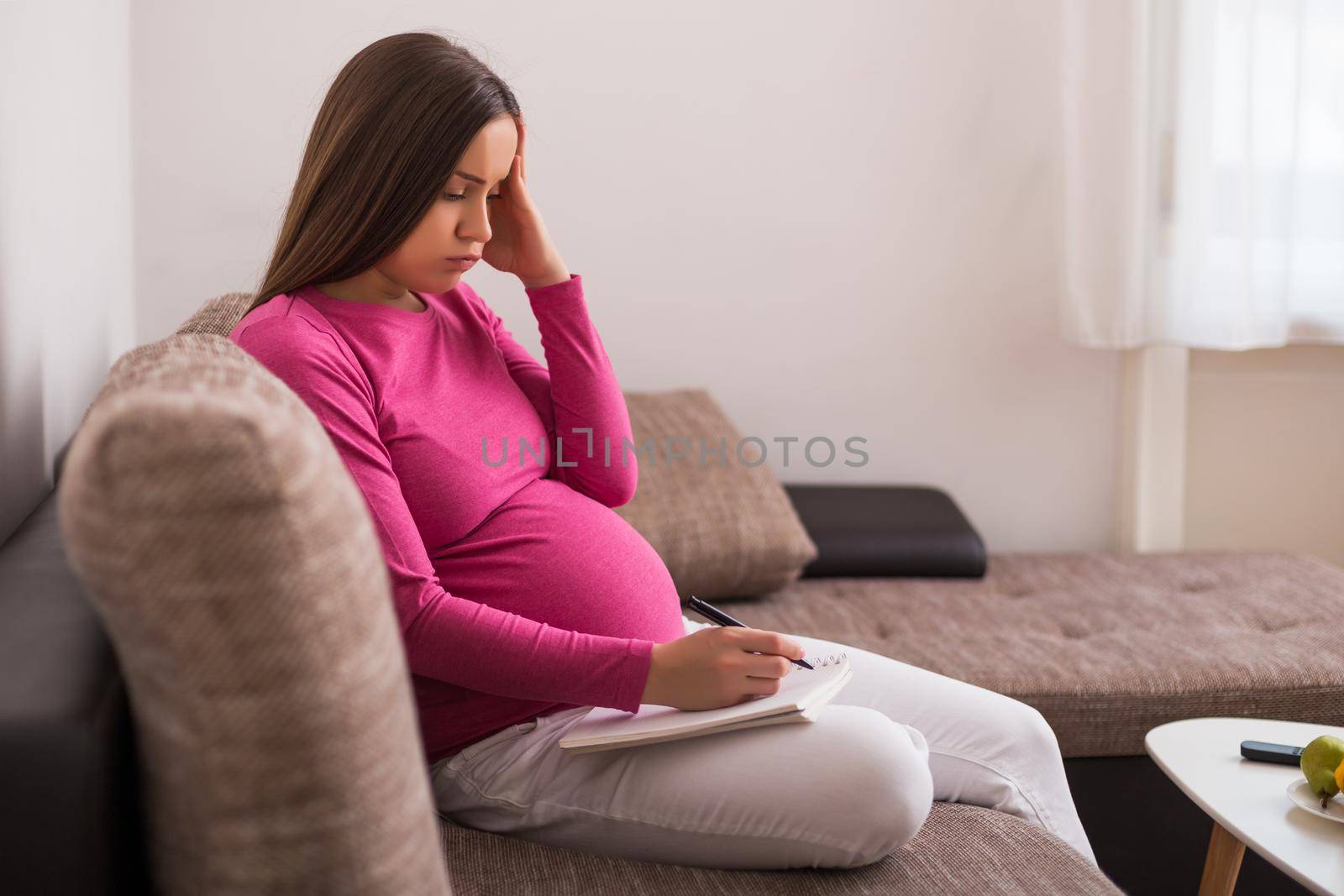 Worried pregnant woman writing to do list in notebook while spending time at her home.
