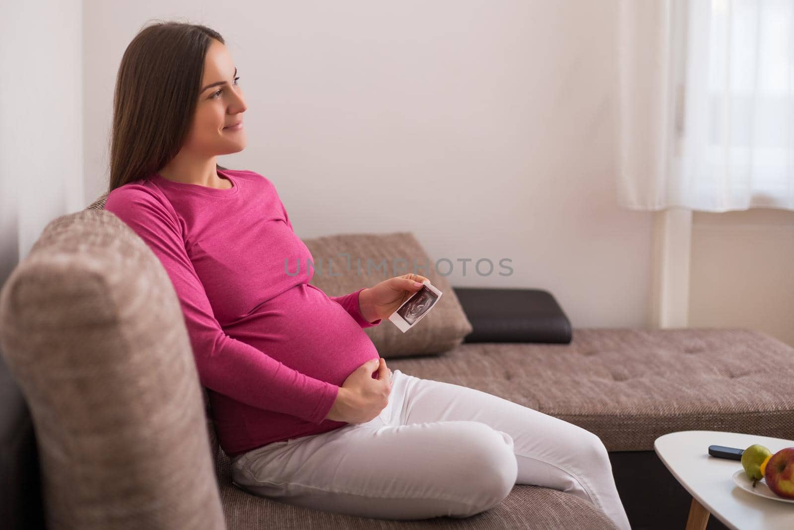 Pregnant woman sitting on the sofa and holding ultrasound picture.