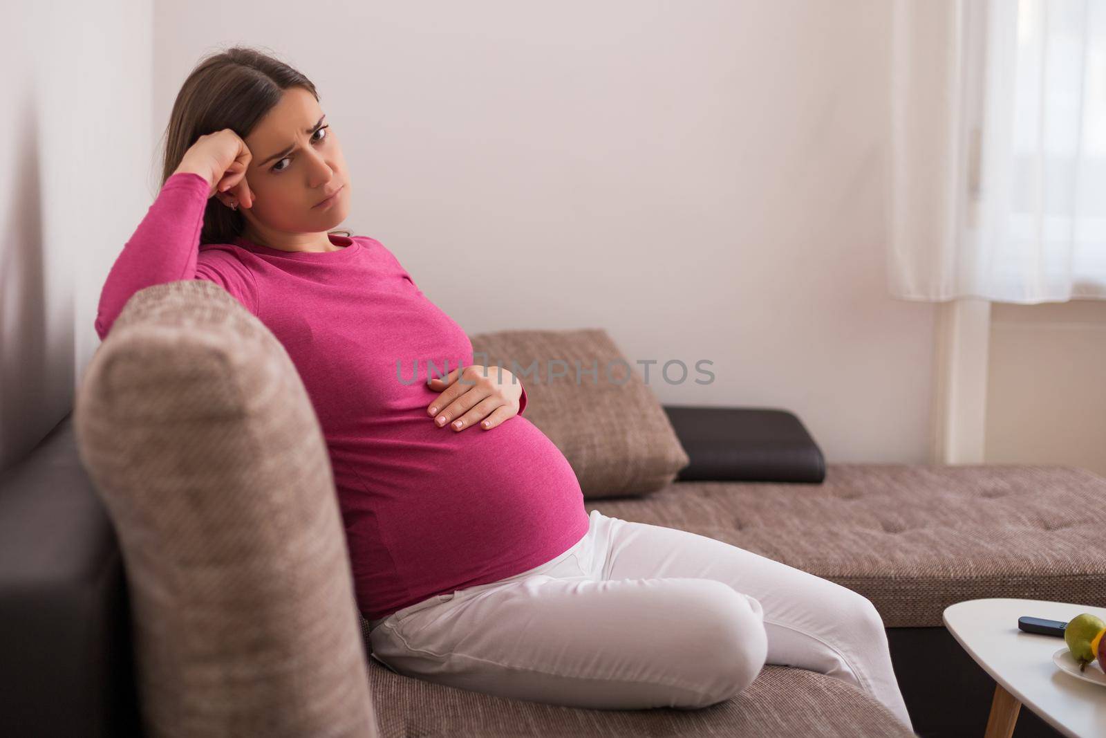 Sad pregnant woman tired of pregnancy is sitting on sofa at her home.