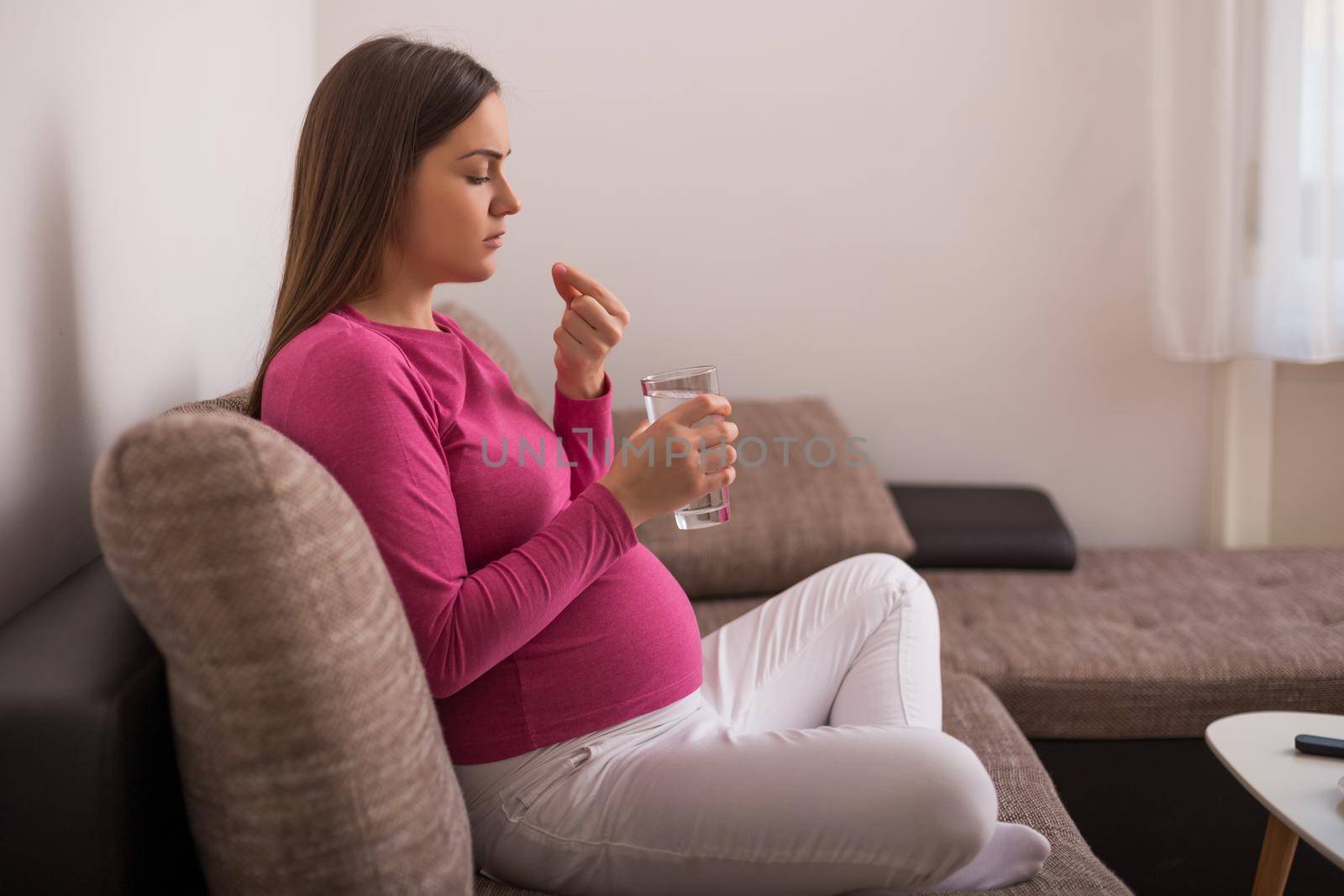 Sad pregnant woman drinking pill while sitting on sofa at her home.