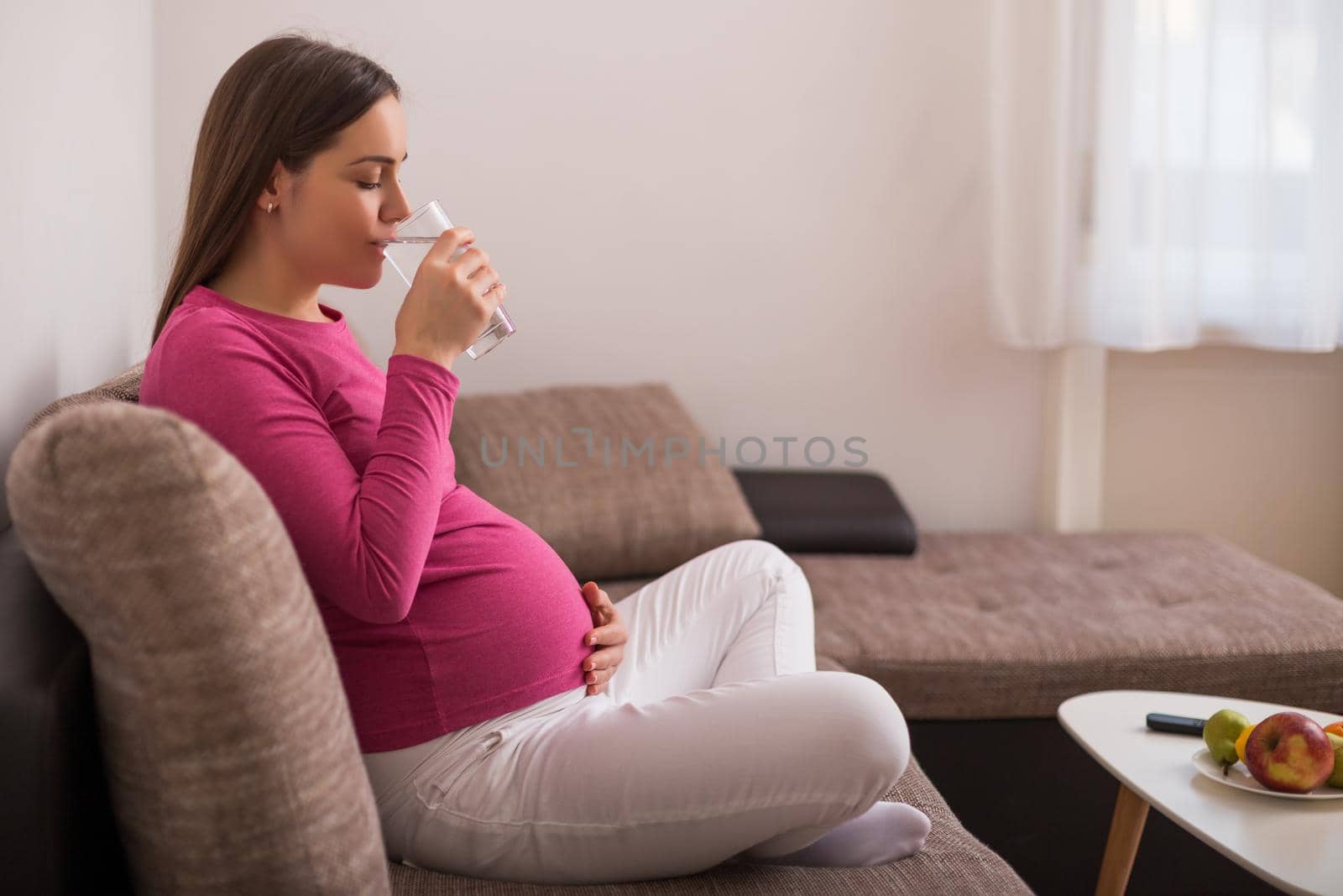 Beautiful pregnant woman enjoys drinking water while  resting on sofa at her home.