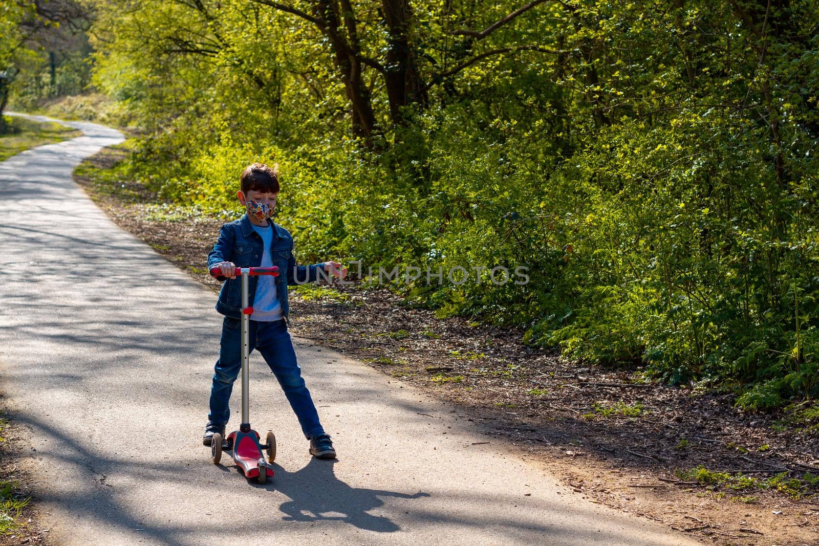 Full-length shot of a boy wearing jeans, a denim jacket and a face mask playing with a scooter in a park on a sunny day