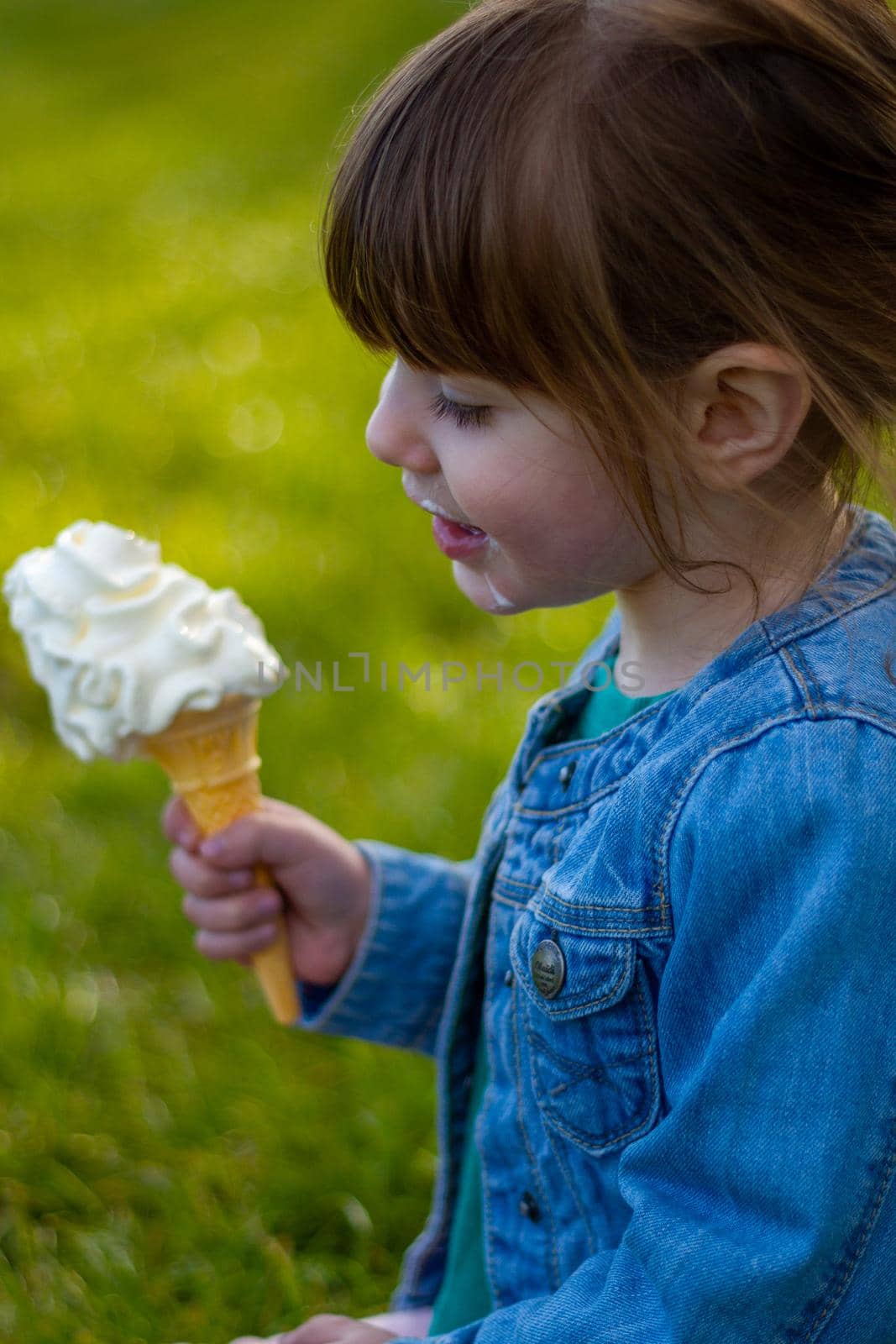 Close-up of a cute, brown-haired, blue-eyed girl, wearing a blue denim jacket eating an ice cream on the grass on a sunny day