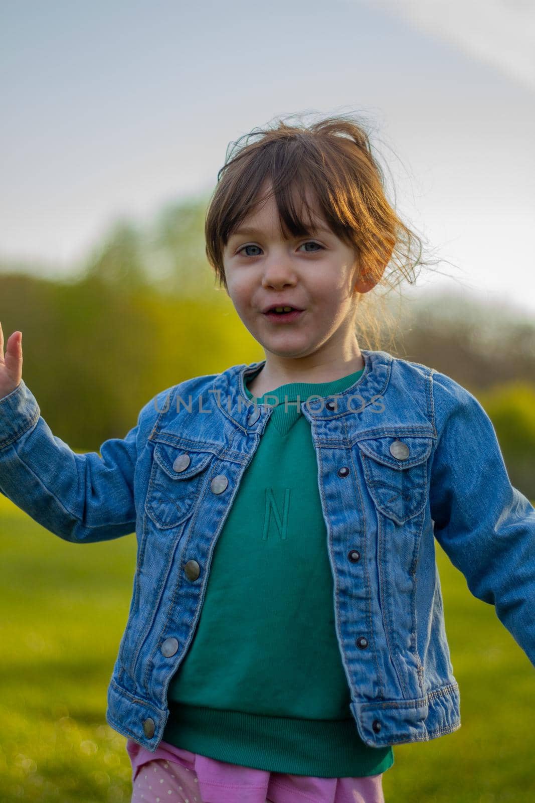Portrait of a cute, brown-haired, blue-eyed baby girl wearing a blue jacket and green jumper running in a park on a sunny day