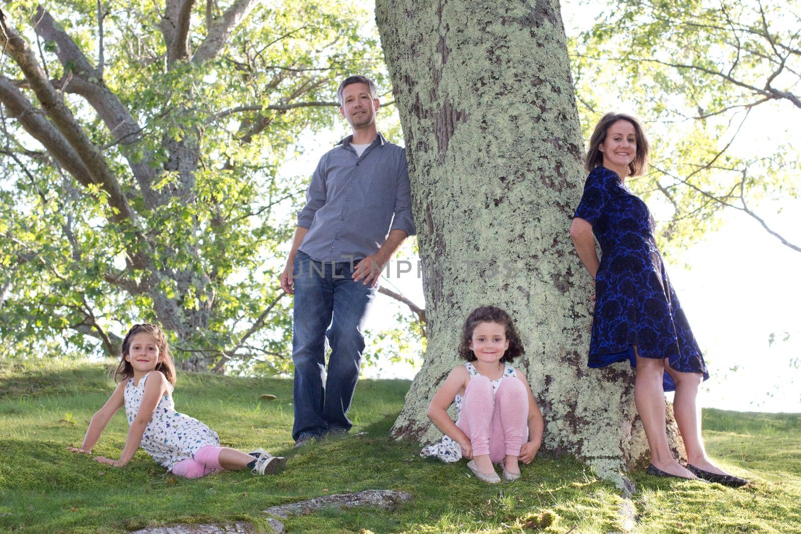 a mother, father and their two daughters casually hang out by a beautiful old tree outdoors smiling