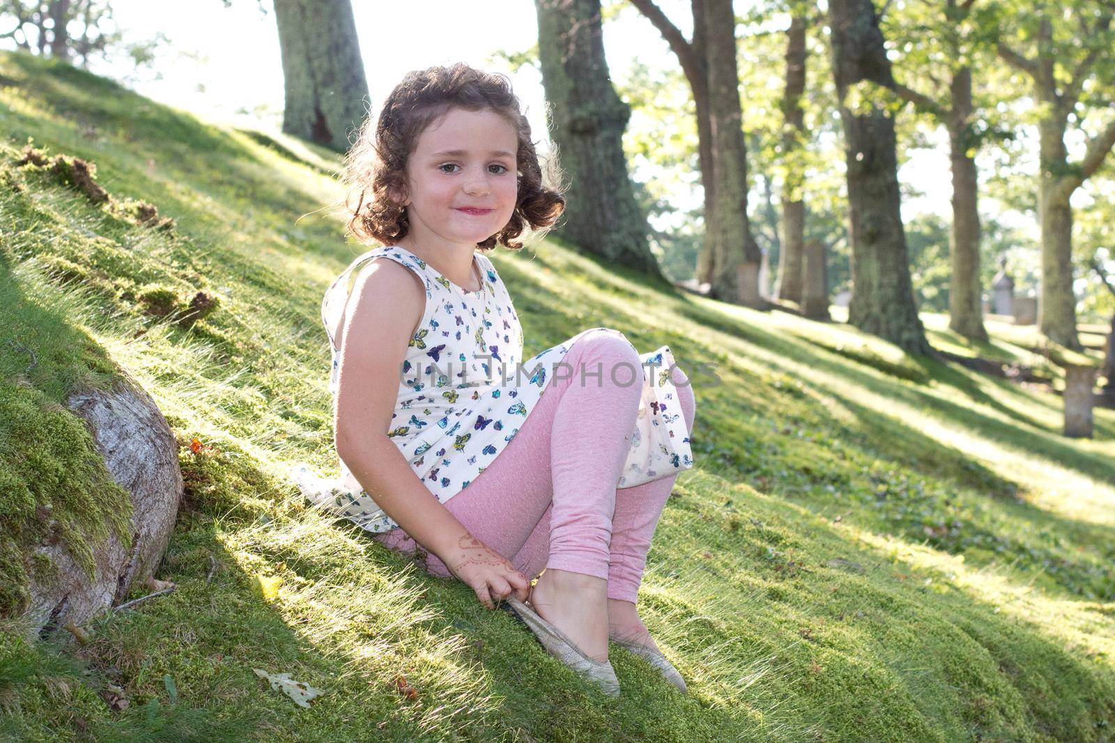  little curly haired girl with an adorable smile sits outside in the summer