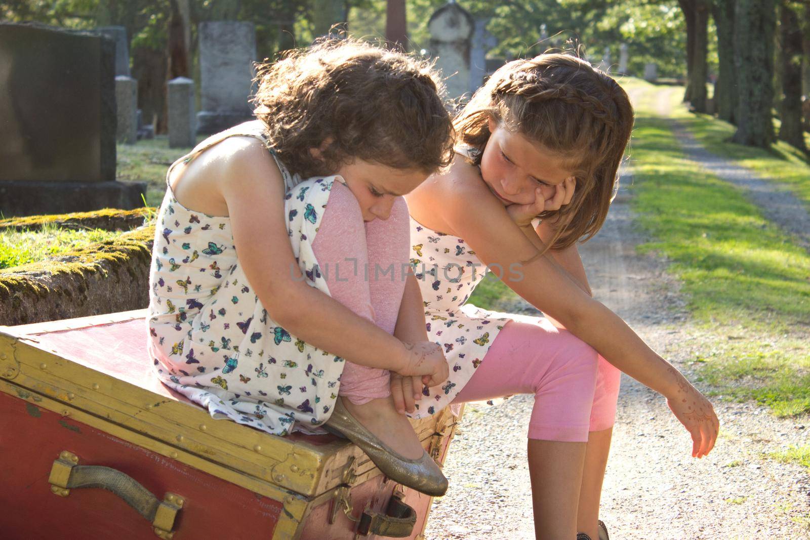 two sad brown haired girls sit on a red box in the cemetery with sad expressions