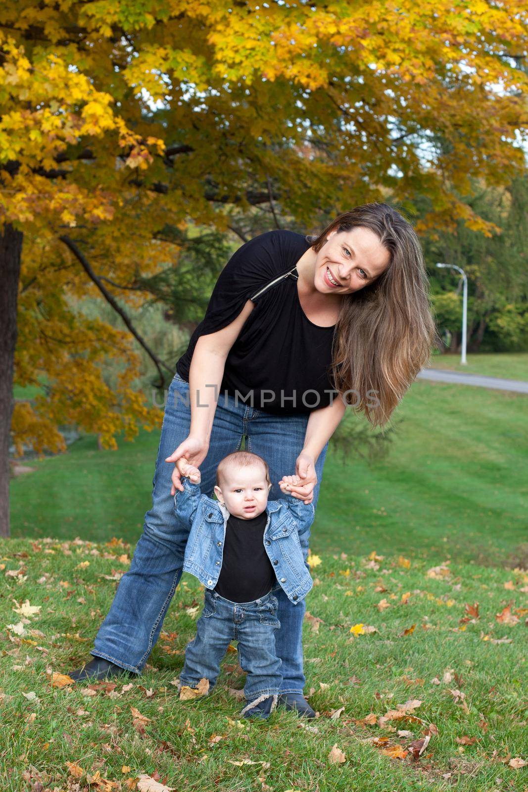 Portrait of mother and baby daughter in the park beside trees with Autumn leaves. The mother is holding baby's hands teaching her to walk.  