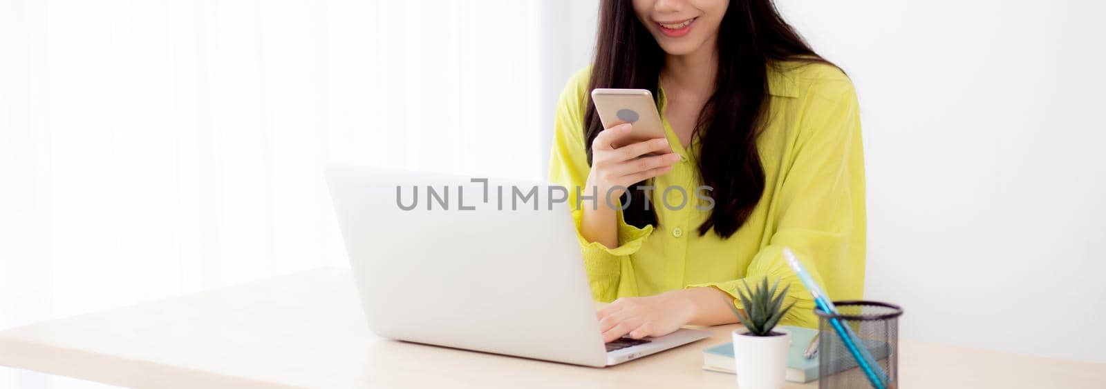 Young asian businesswoman working on laptop computer online on desk at home office, freelance woman looking message on smart phone and typing on notebook on table, business and communication concept.