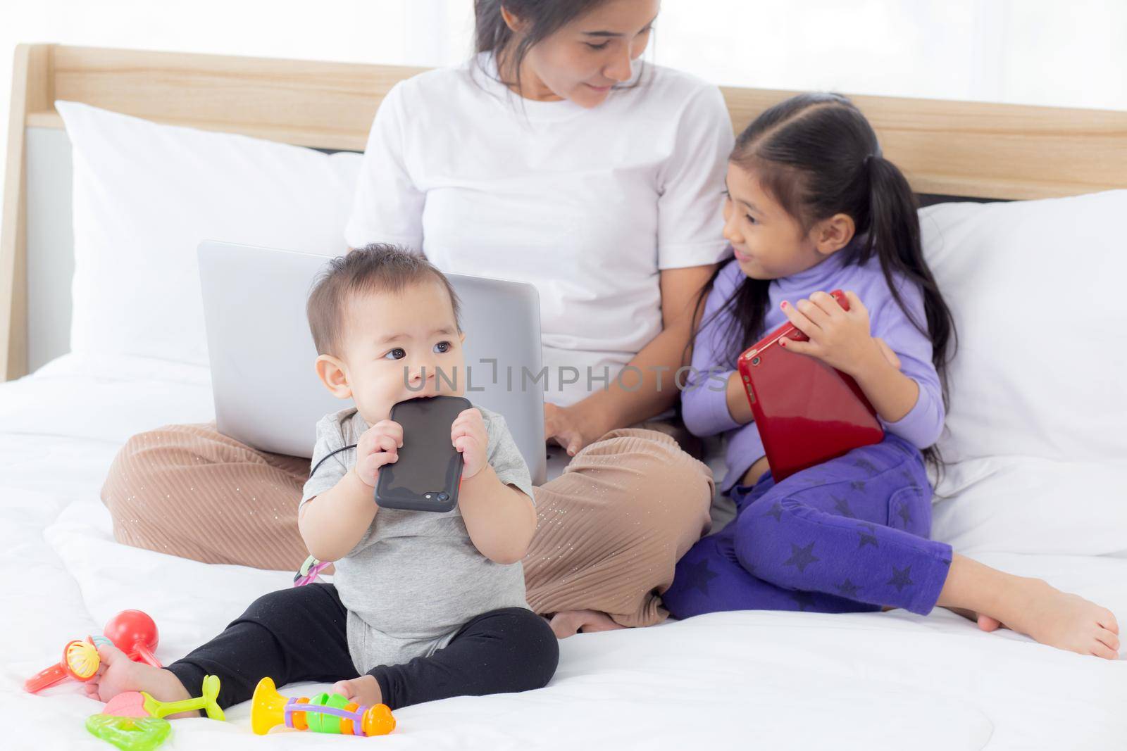 Young asian mother working at home online to internet with laptop computer and daughter looking tablet on bed at bedroom, business woman is freelance and children learning, new normal, family concept.