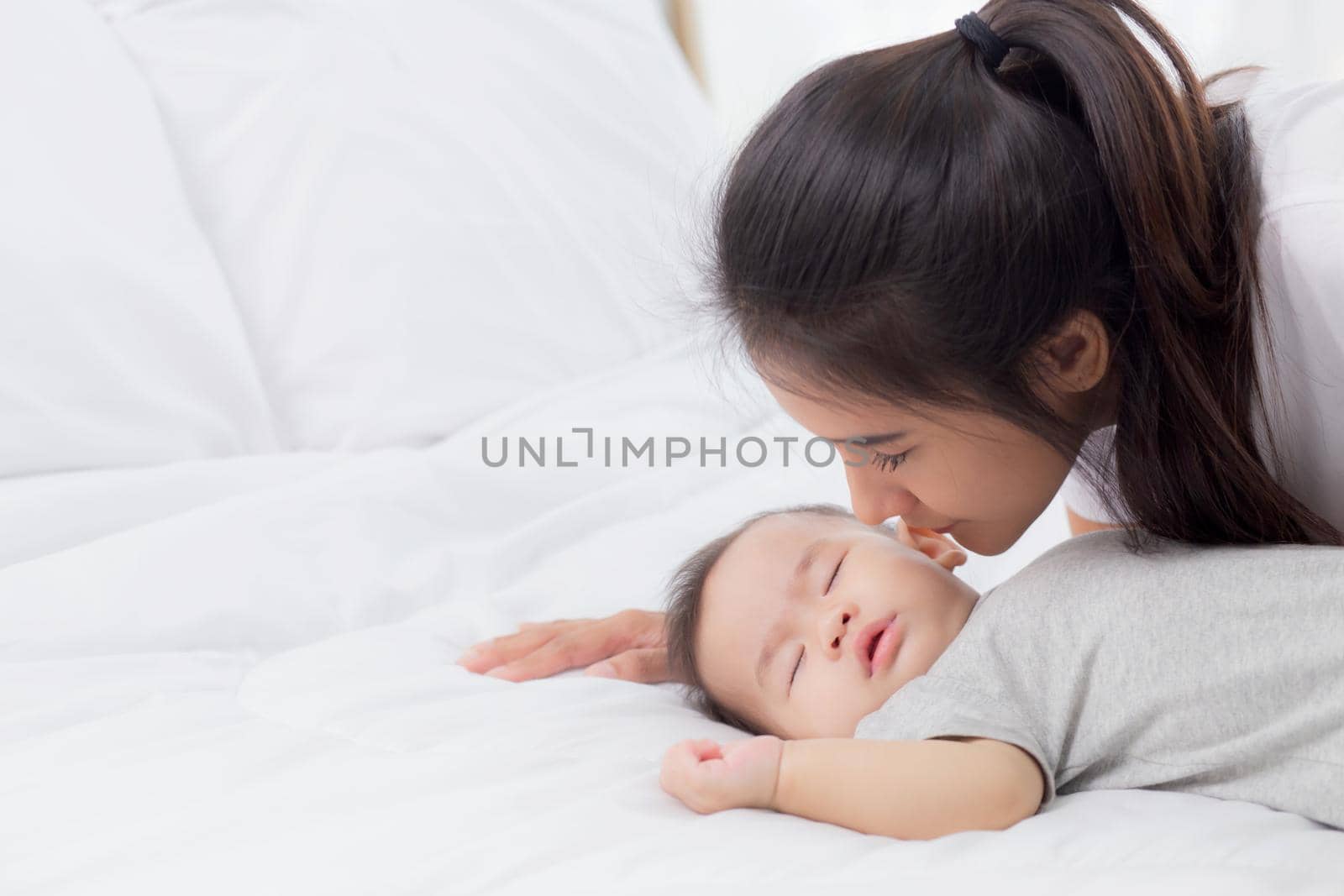 Young asian mother kiss cheek of little baby girl with tender on bed in the bedroom, mom love newborn and care, mother with expression with child together, parent and daughter, family concept.