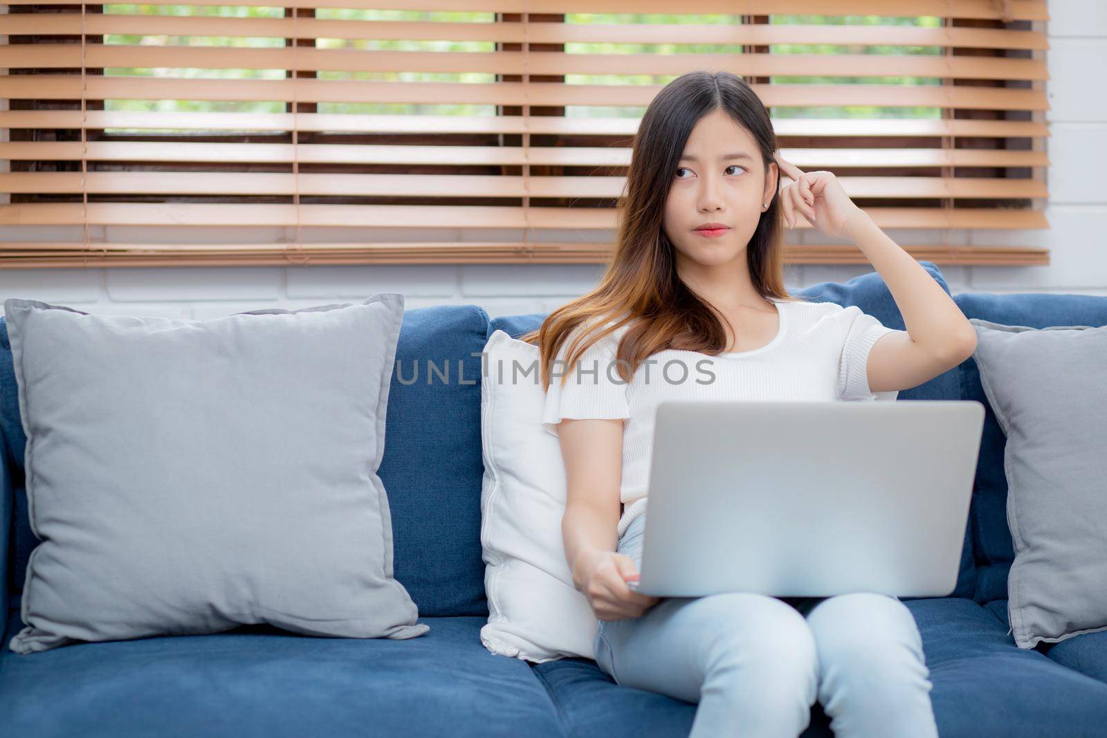 Beautiful young asian woman working on laptop computer and thinking idea on sofa at home, freelance girl sitting on couch using notebook to internet at living room, one person, lifestyle concept.