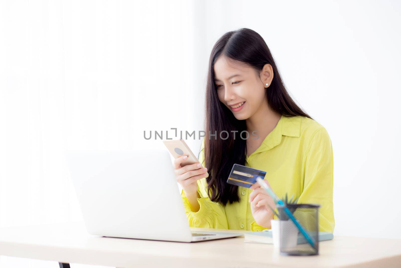 Young asian business woman using smart phone and holding credit card while online shopping and payment online with laptop computer on desk at home, female holding debit card, communication concept.