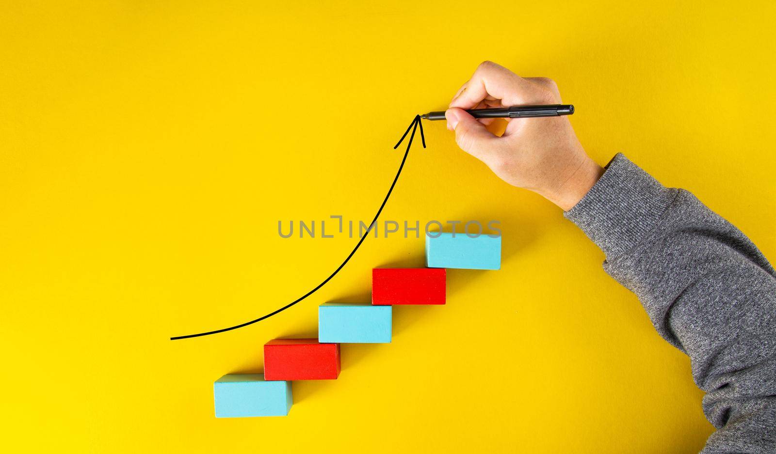 Male hand drawing an upward pointing arrow on top of growing graph made of wooden blocks over yellow background