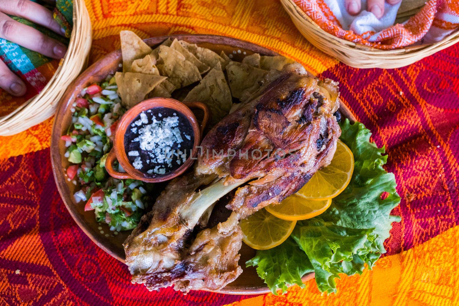 Top view of delicious roasted pork with lemon slices, lettuce, tortilla chips, and refried beans on colorful tablecloth. Tasty meat and accompaniments above orange and red table. Mexican style meal