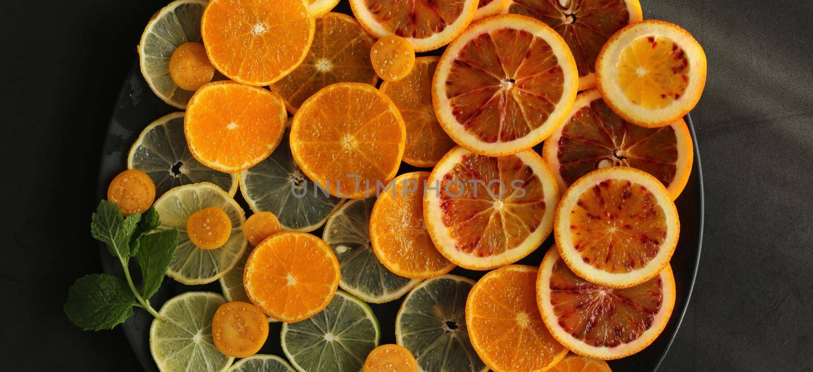 Fresh citruses on round plate on dark black stone background, flat lay. Oranges, clementine, mandarins, limes. Top view. Dramatic kitchen