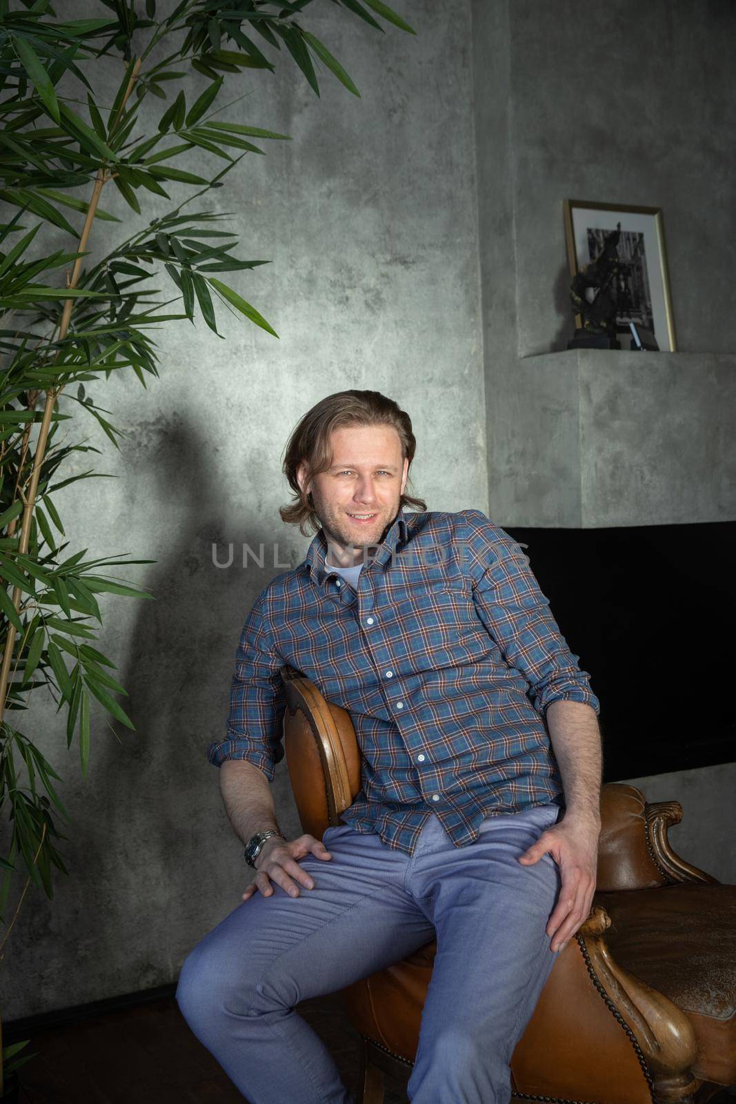 The handsome stylish young man sits on an expensive leather armchair relaxed, long curly hair, he is dressed in a blue shirt and trousers, he is looking at the camera by vladimirdrozdin