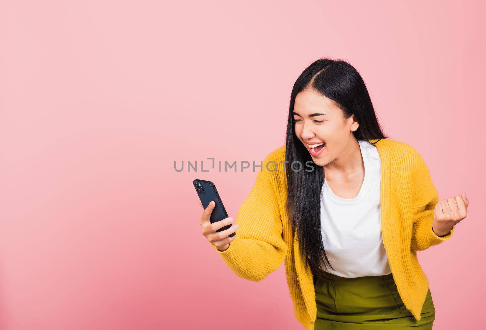 Happy Asian portrait beautiful cute young woman teen smiling excited   using mobile phone say yes! Studio shot isolated on pink background, Thai female surprised make winner gesture on smartphone