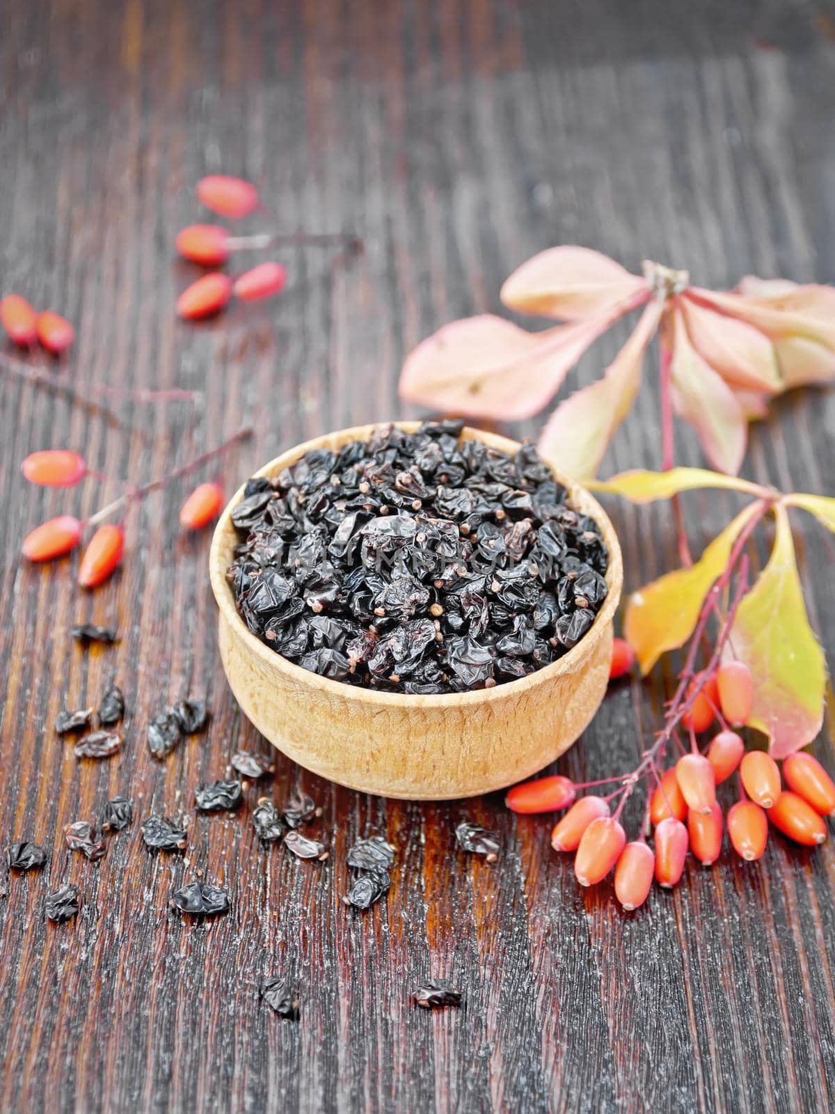 Dried black barberry in a bowl, sprigs with fresh berries and leaves on brown wooden board background