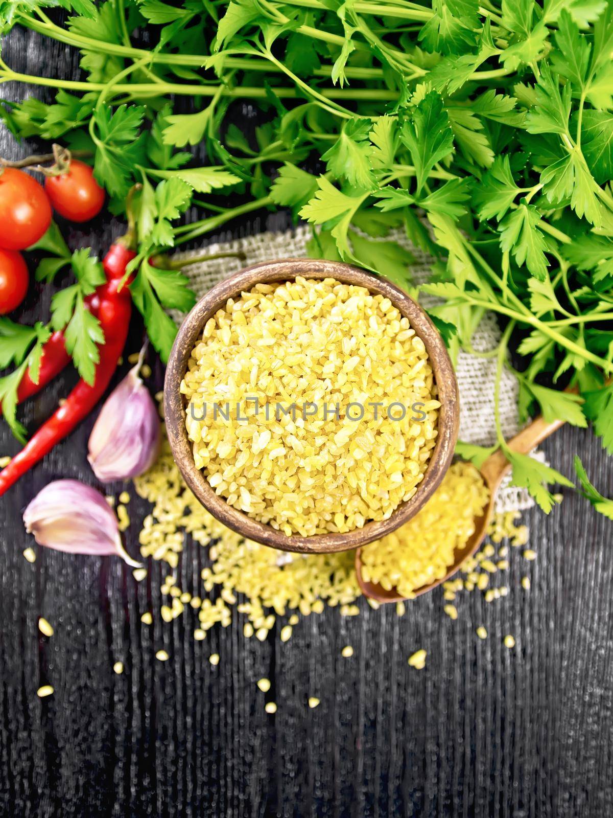 Bulgur groats - steamed wheat grains - in a clay bowl and spoon on sacking, tomatoes, hot peppers, garlic and parsley on wooden board background from above
