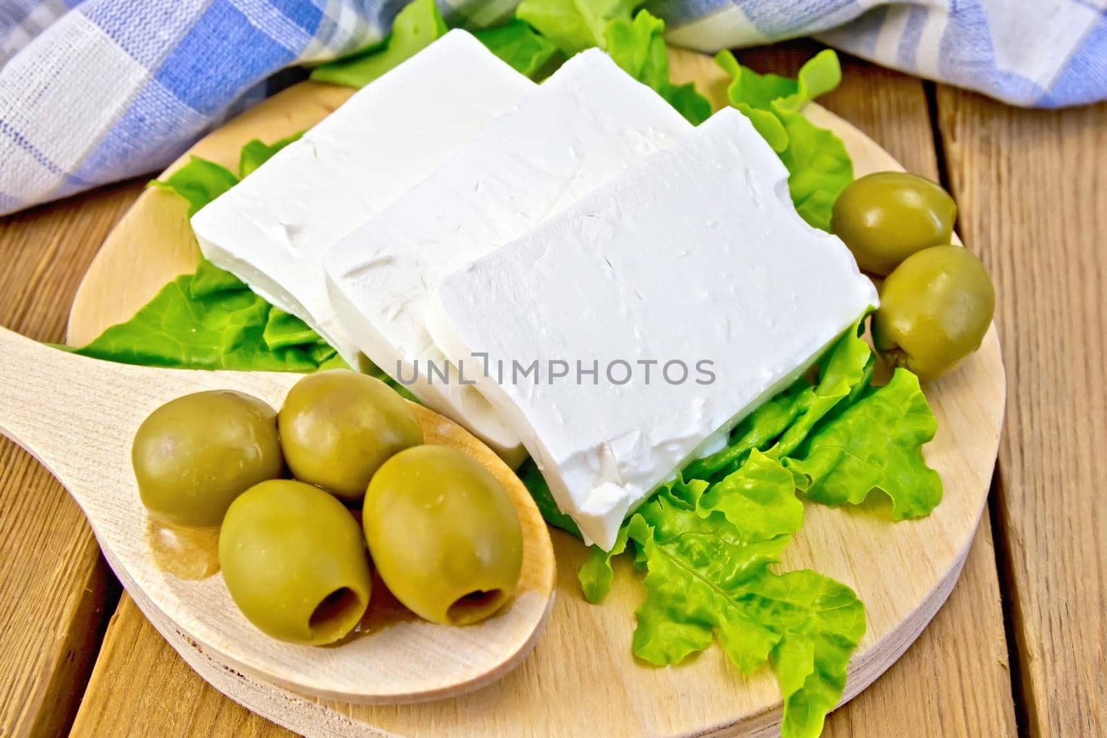 White brine cheese, lettuce, olives, spoon with olives, blue plaid napkin on a wooden boards background