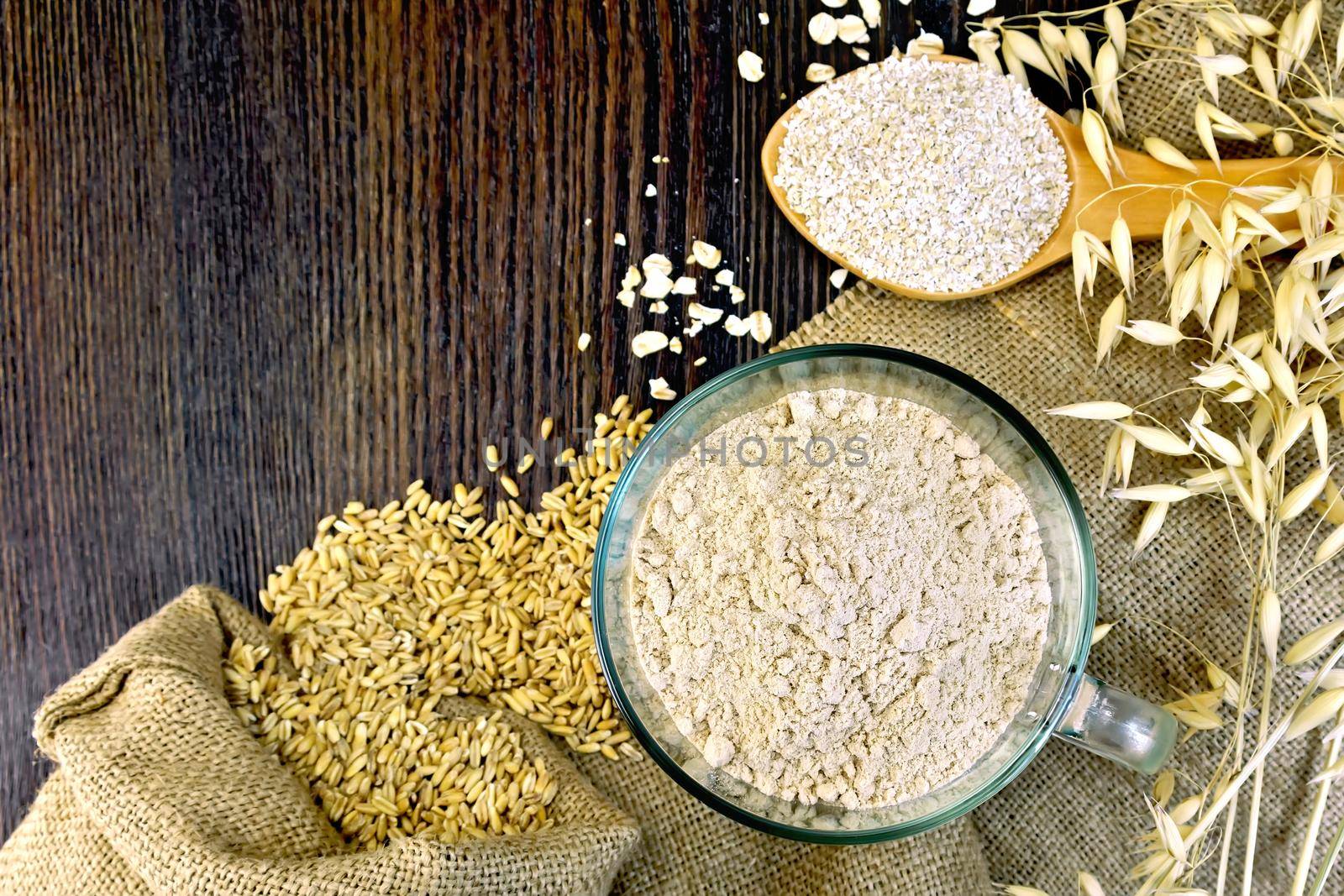 Oat Flour in a glass bowl, bran flakes in a spoon, oats and oatmeal stalks on sackcloth background on wooden board