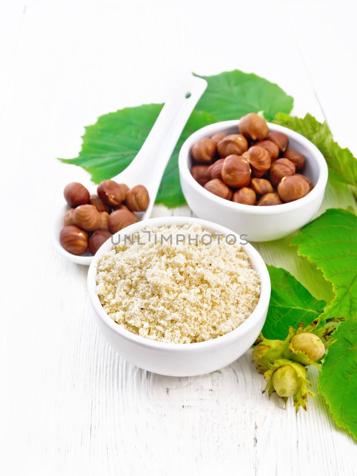Flour and hazelnuts in two bowls, a spoon with peeled walnut kernels and a branch of filbert with leaves on wooden board background