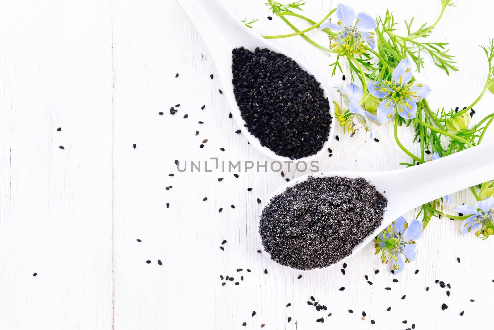 Flour and seeds of Nigella sativa in two spoons, sprigs of kalingini with blue flowers and green leaves on a wooden board background from above
