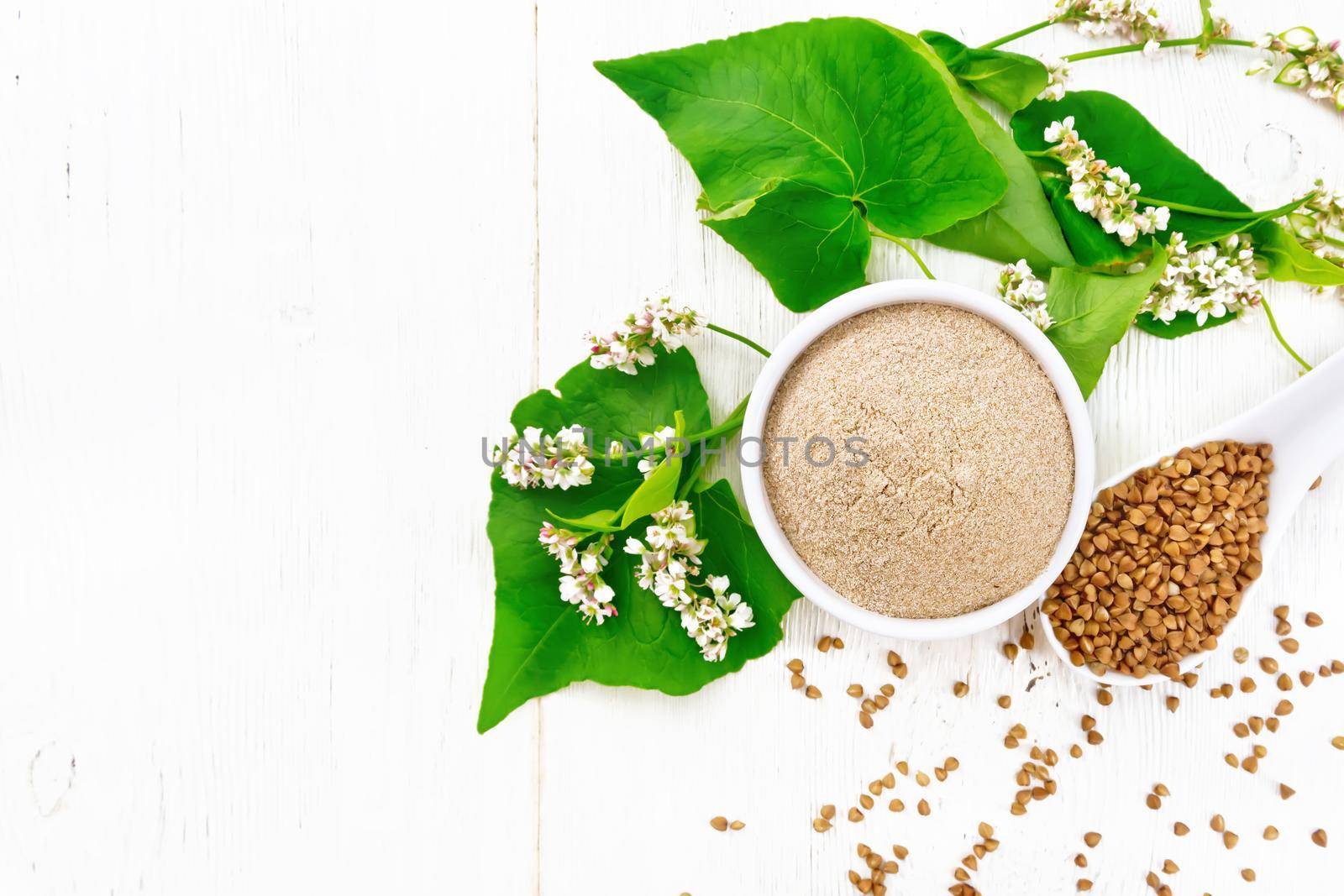 Brown buckwheat flour in a bowl, brown groats in a spoon, flowers and buckwheat leaves on wooden board background from above