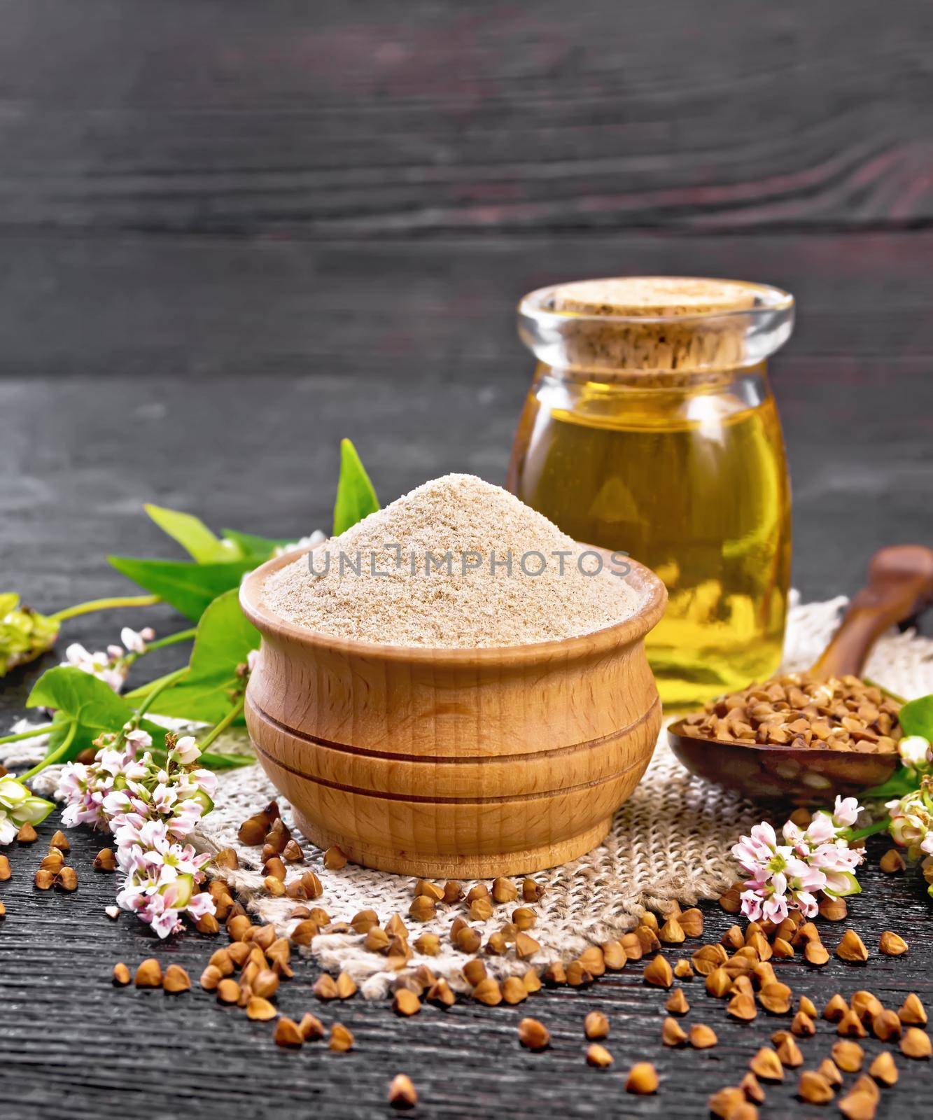 Brown buckwheat flour in a bowl, brown groats in a spoon, buckwheat flowers and leaves, oil in a glass jar on burlap on the background of dark wooden board