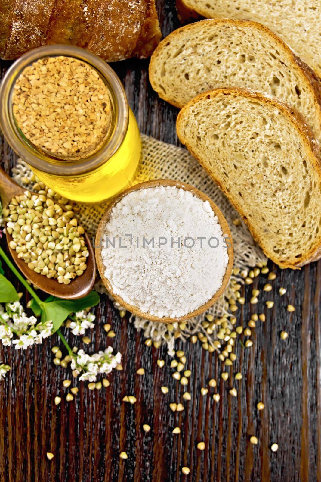 Buckwheat flour from green cereals in a bowl on sacking, groats in spoon and on a table, oil in glass jar, bread, fresh flowers and buckwheat leaves on background of dark wooden board from above