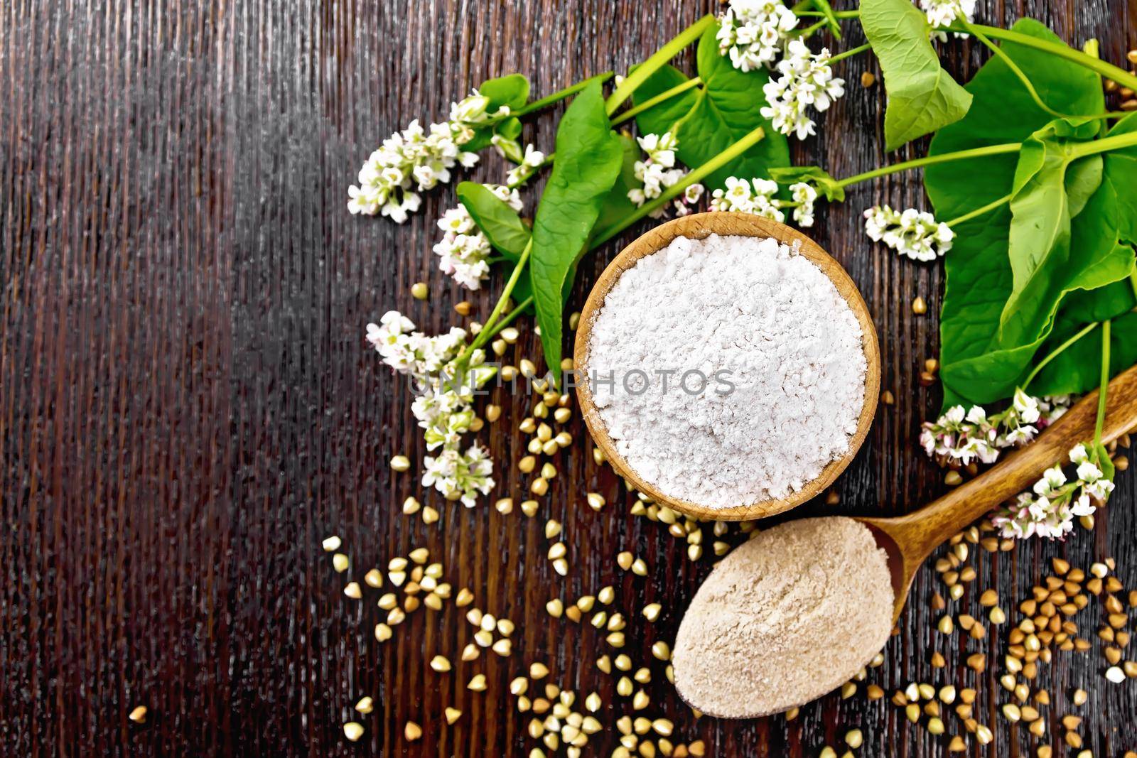 Buckwheat flour from green cereals in a bowl, buckwheat flour from brown groats in a spoon, flowers and leaves on the background of a wooden board from above