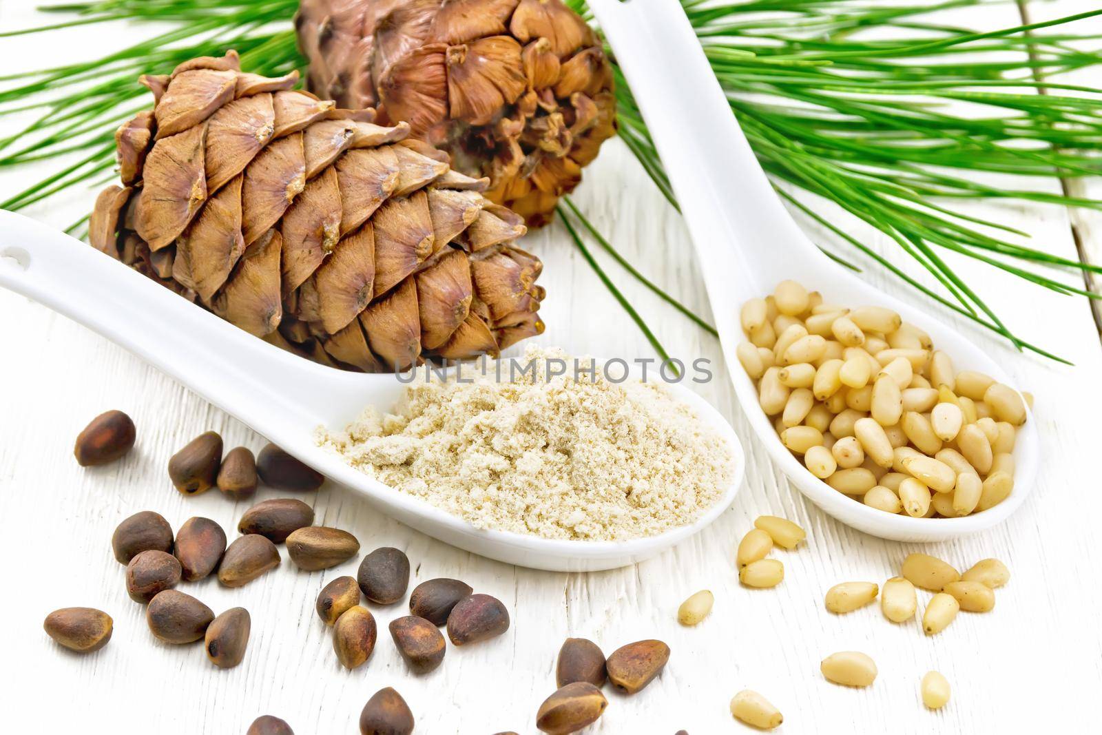 Cedar flour and peeled nuts in two spoons, two cones, a green cedar branch on background of a light wooden board