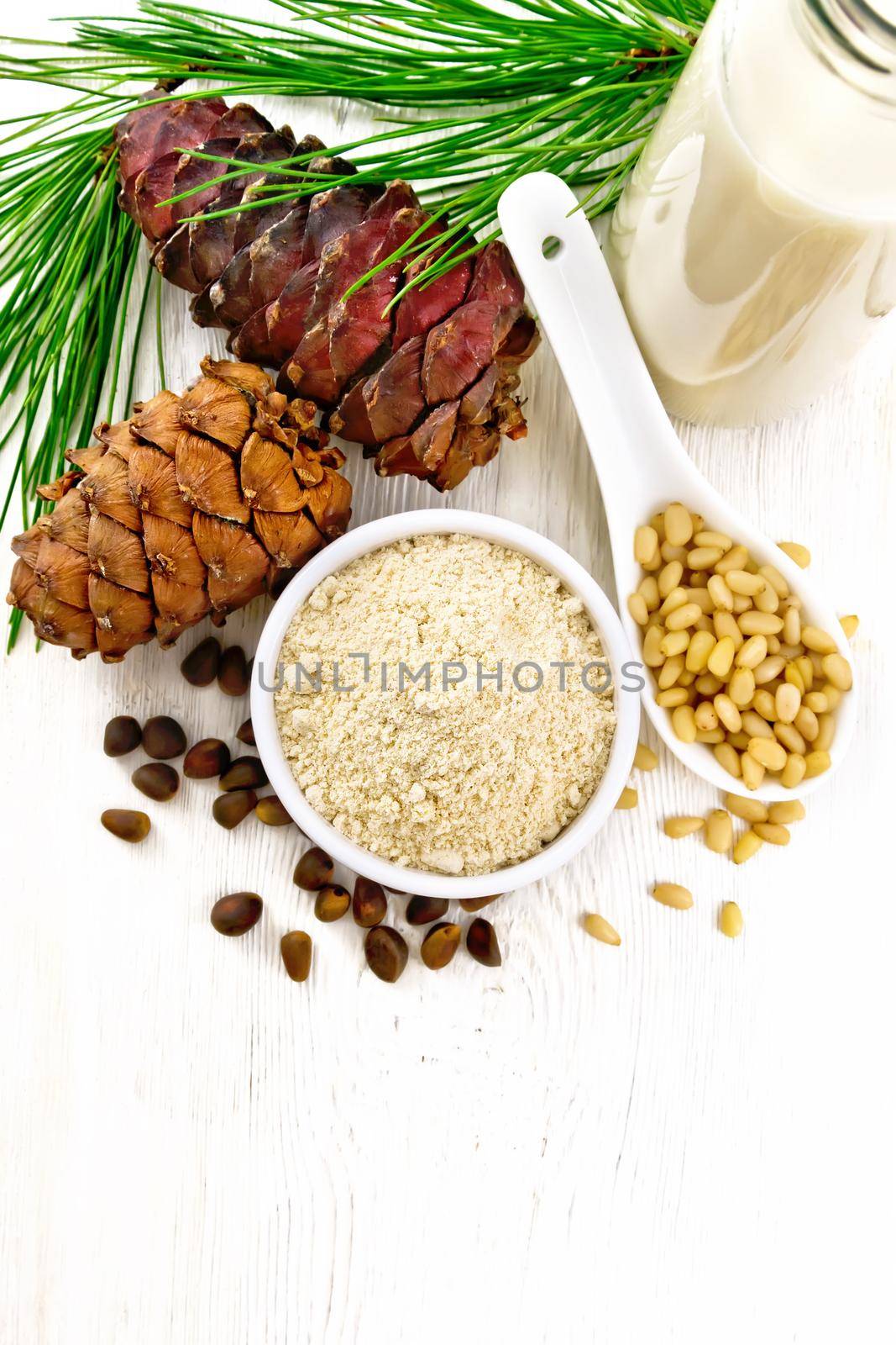 Cedar flour in a bowl, nuts and cones, spoon with peeled nuts, green pine branch and cedar milk in a bottle on white wooden board background from above