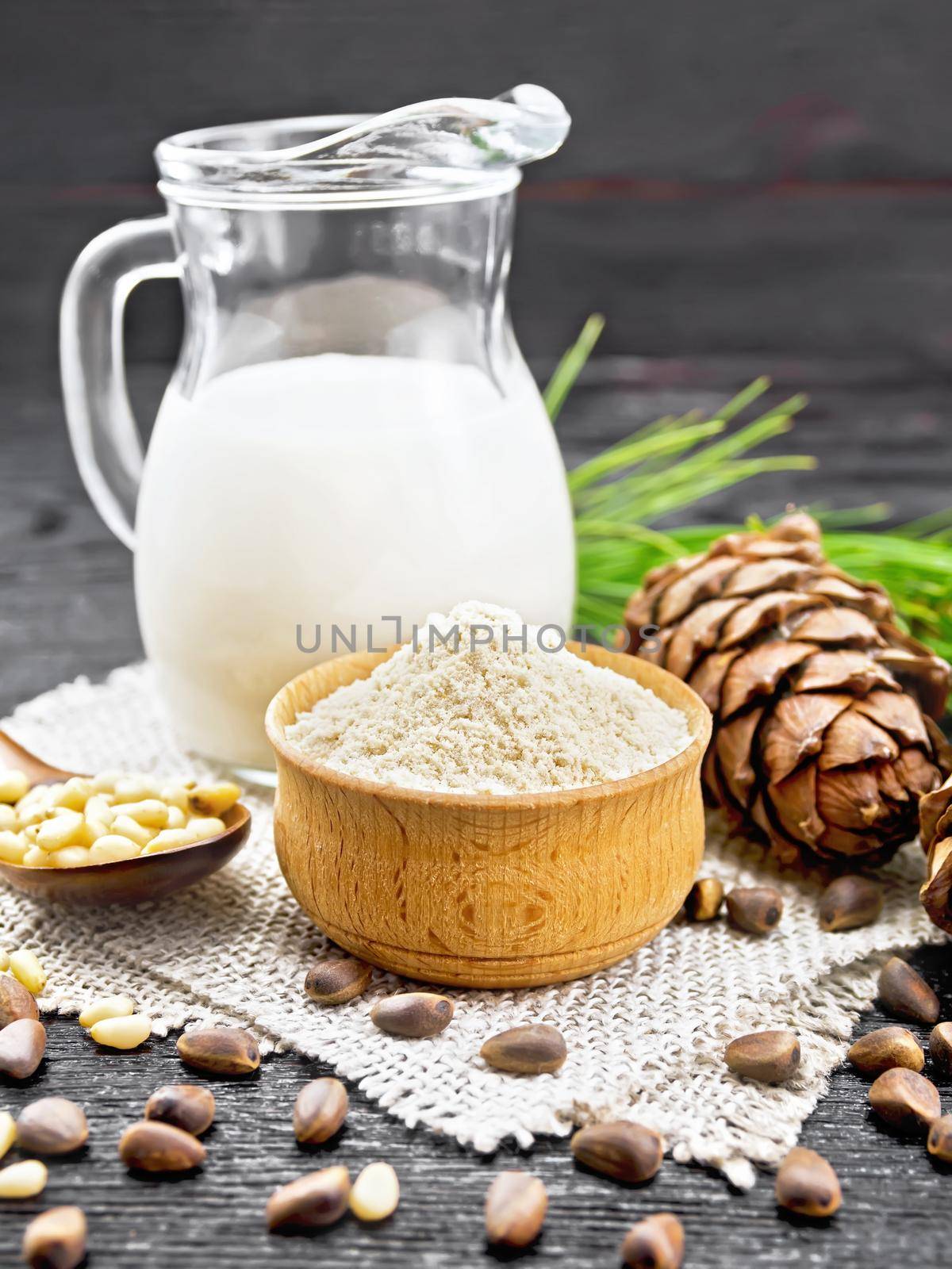 Cedar flour in a bowl, nuts and two cones, spoon with peeled nuts on burlap napkin, green pine branch and cedar milk in a jug on black wooden board background
