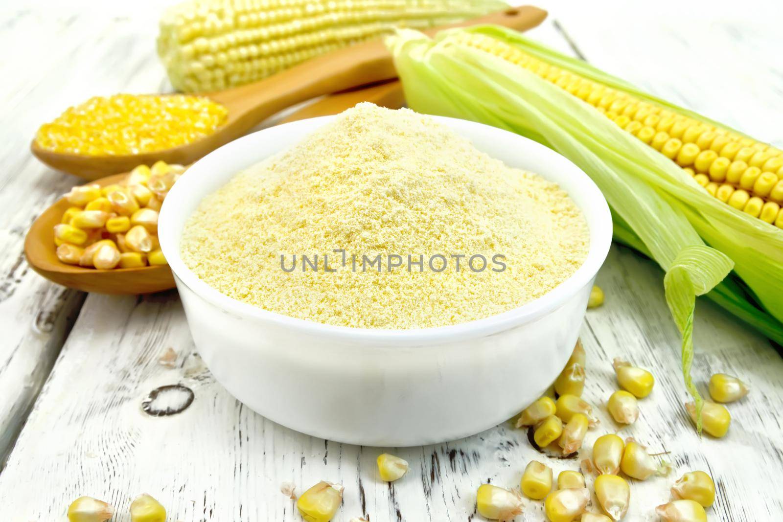 Flour corn in a bowl, groats and grain maize in the spoons, cobs on the background light wooden boards