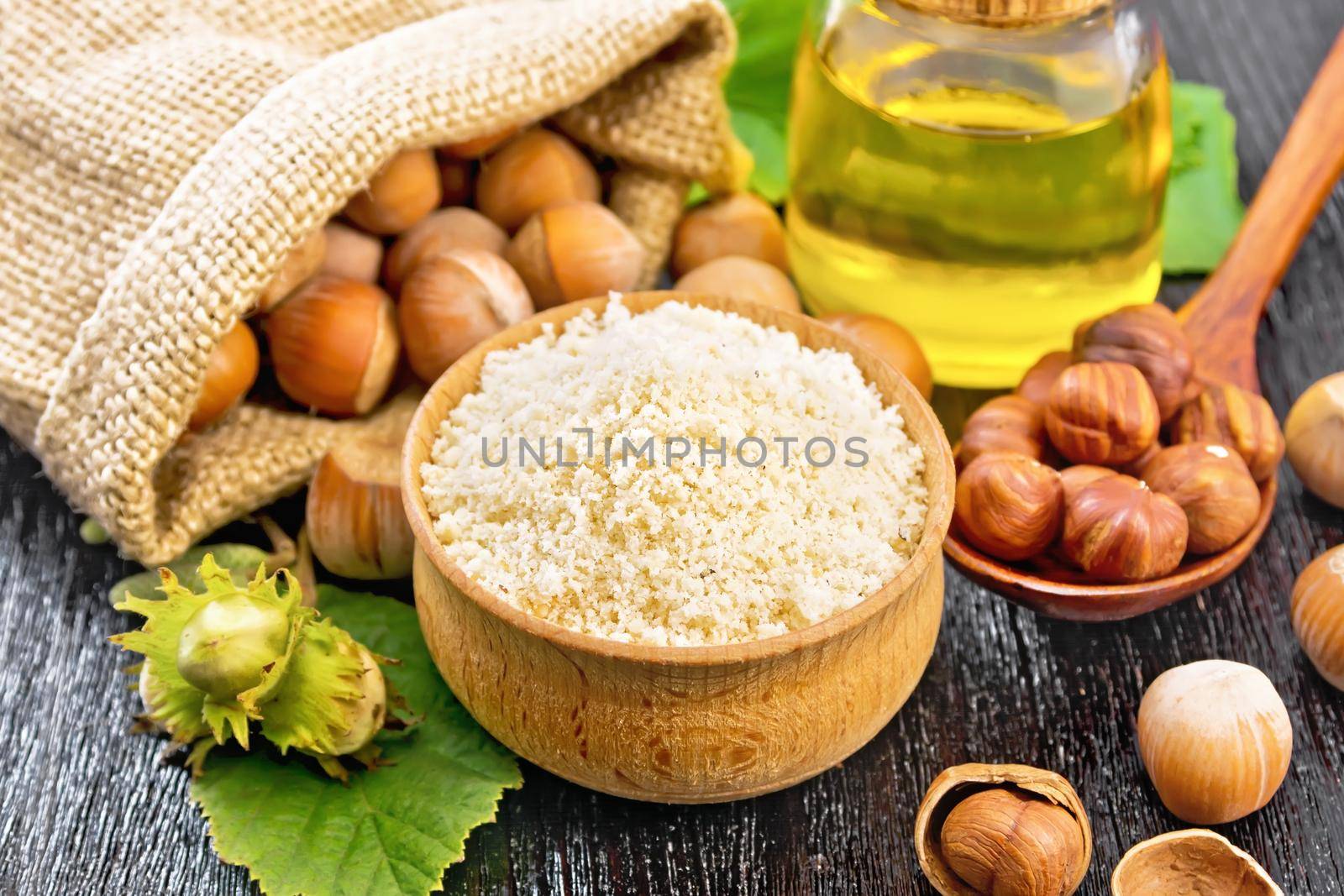 Hazelnut flour in a bowl, nuts in bag, a spoon, oil in glass jar and filbert branch with green leaves on wooden board background