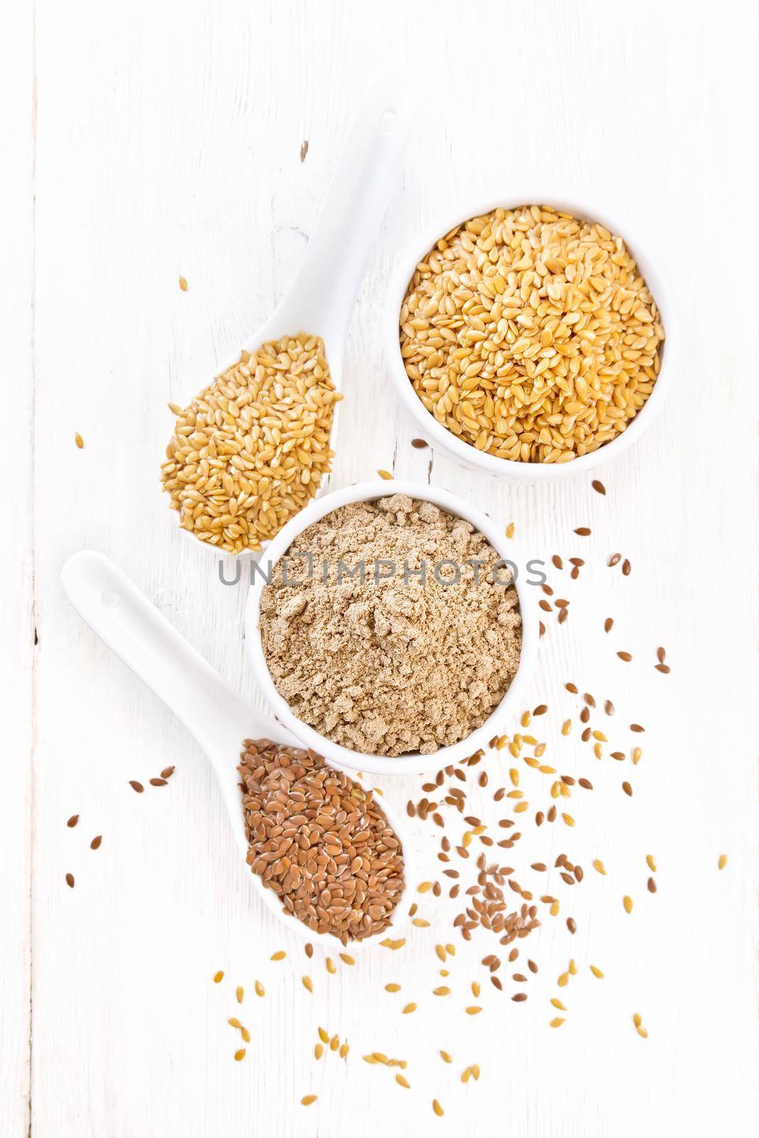 Flaxseed flour in a bowl, white and brown linen seeds in two spoons and on table on wooden board background from above