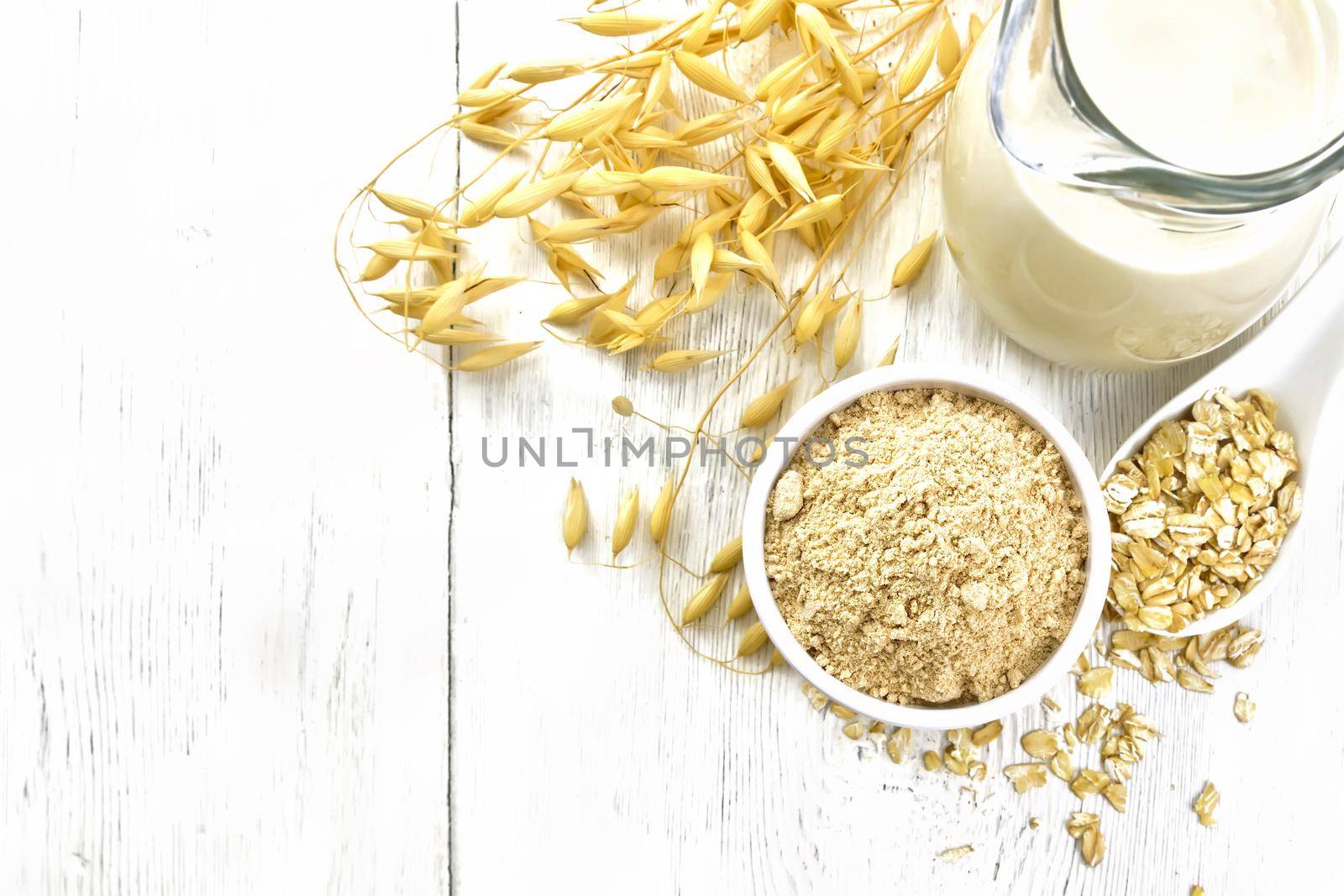 Flour oat in bowl, milk in a jug, oatmeal in spoon, oaten stalks on wooden board background from above