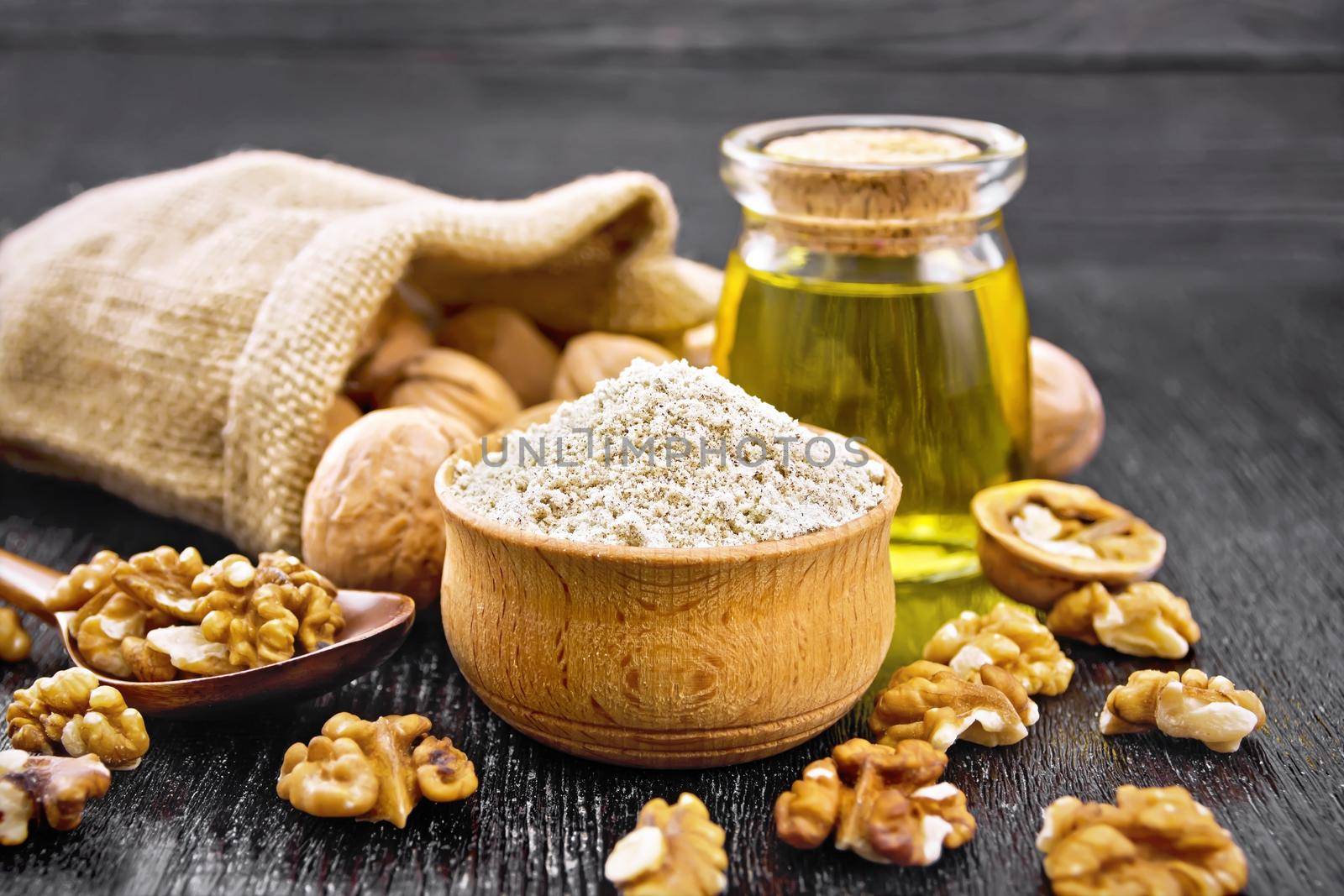 Walnut flour in bowl, nuts on a table, in a spoon and in a bag, oil in glass jar on wooden board background
