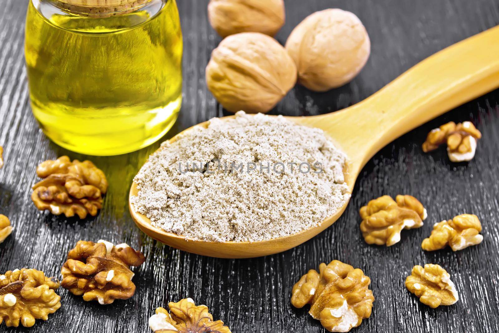 Walnut flour in a spoon, nuts on the table and oil in a glass jar on background of dark wooden board